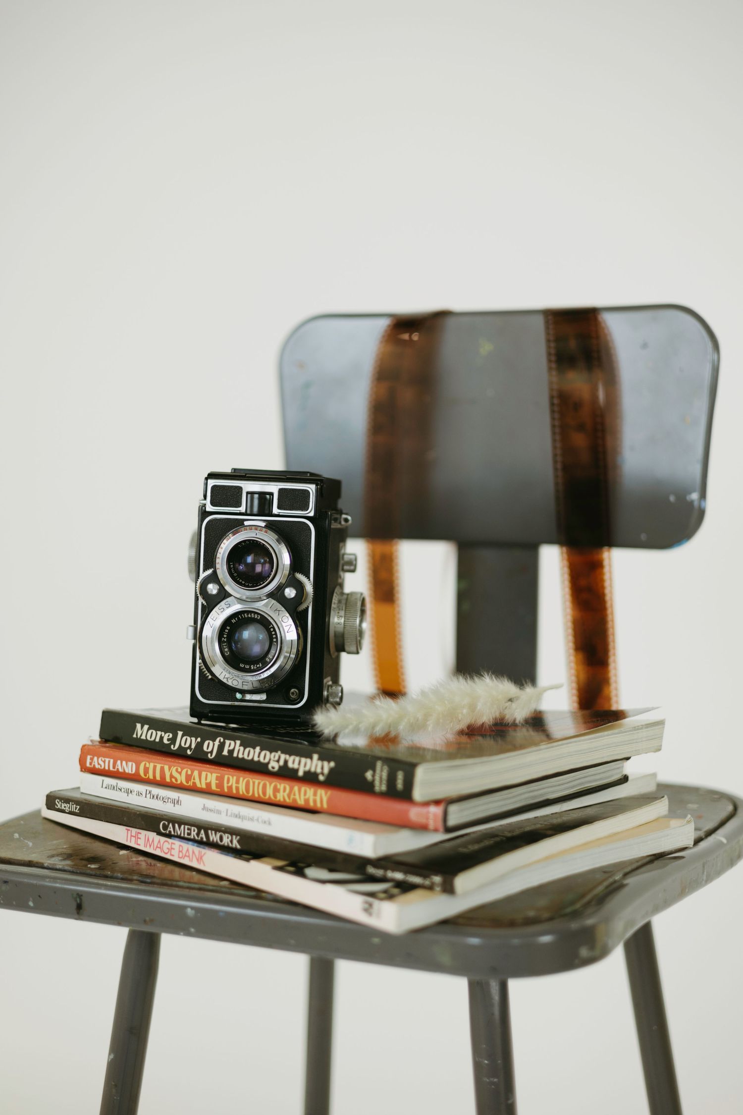 Vintage twin-lens reflex camera and photography books stacked on a metal stool against a light gray background.