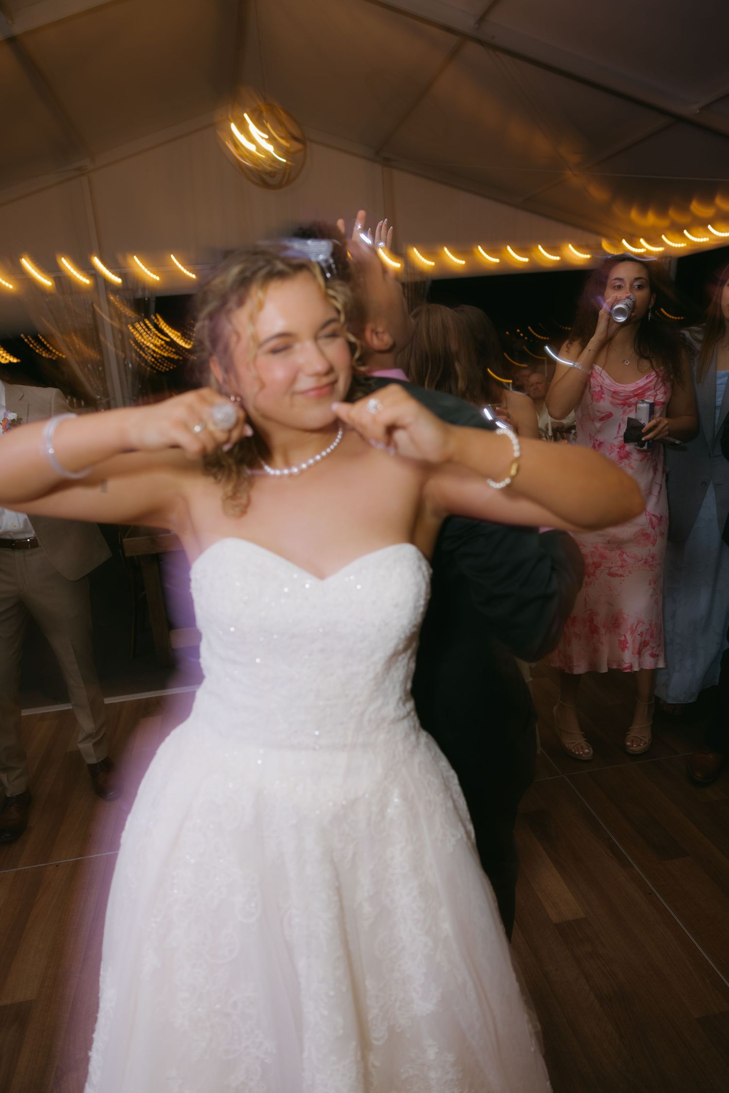 Bride in white strapless ballgown dancing joyfully at wedding reception under warm string lights.