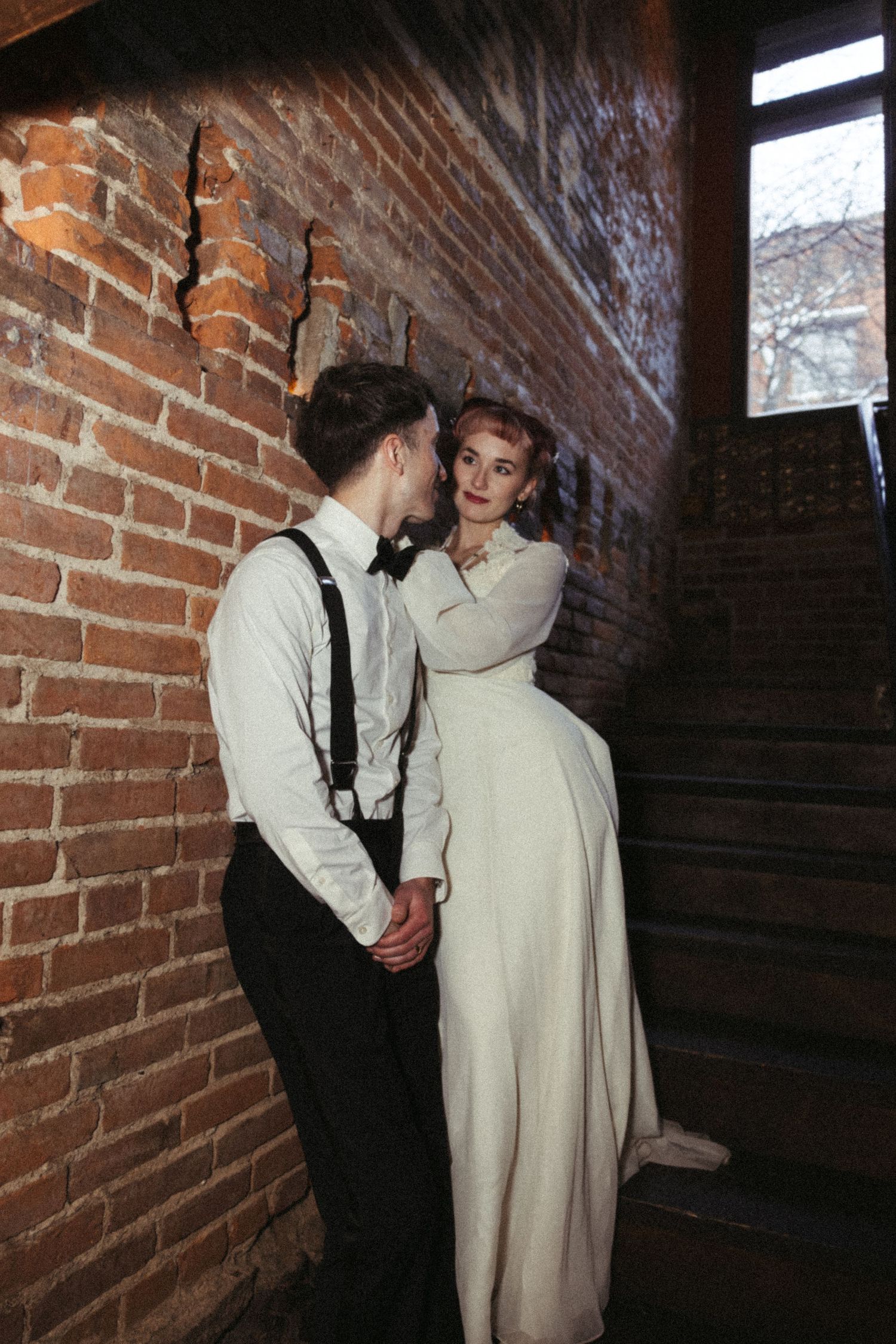 A romantic vintage-style couple embraces on brick stairwell, with the person in a cream colored dress leaning against the wall.