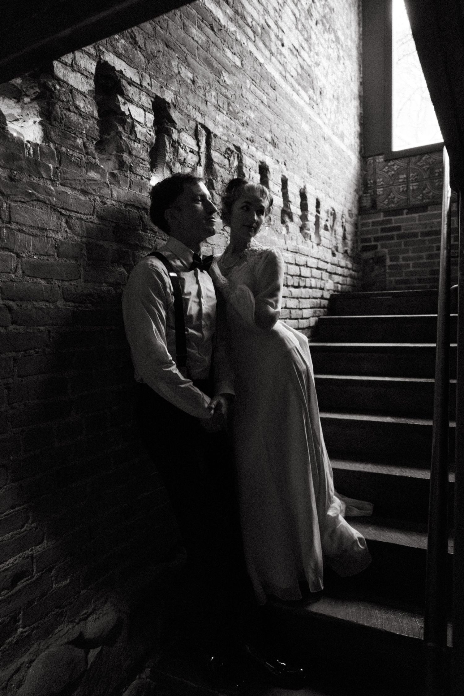 A romantic black and white photograph of a couple in formal attire standing in a dimly lit brick stairwell.