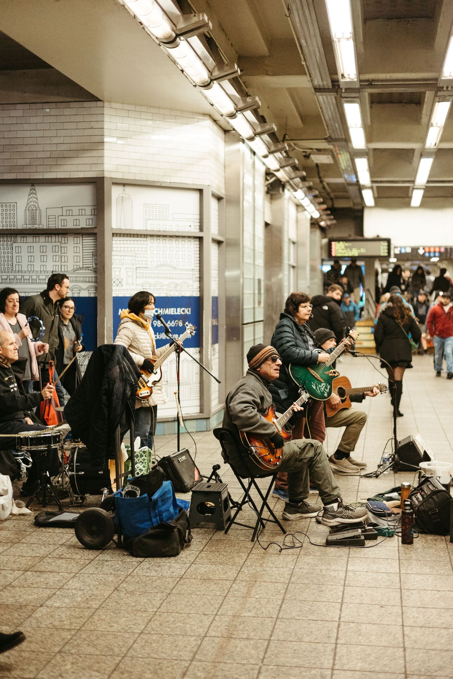 Musicians perform in New York City subway station corridor with instruments and equipment set up along tiled wall.