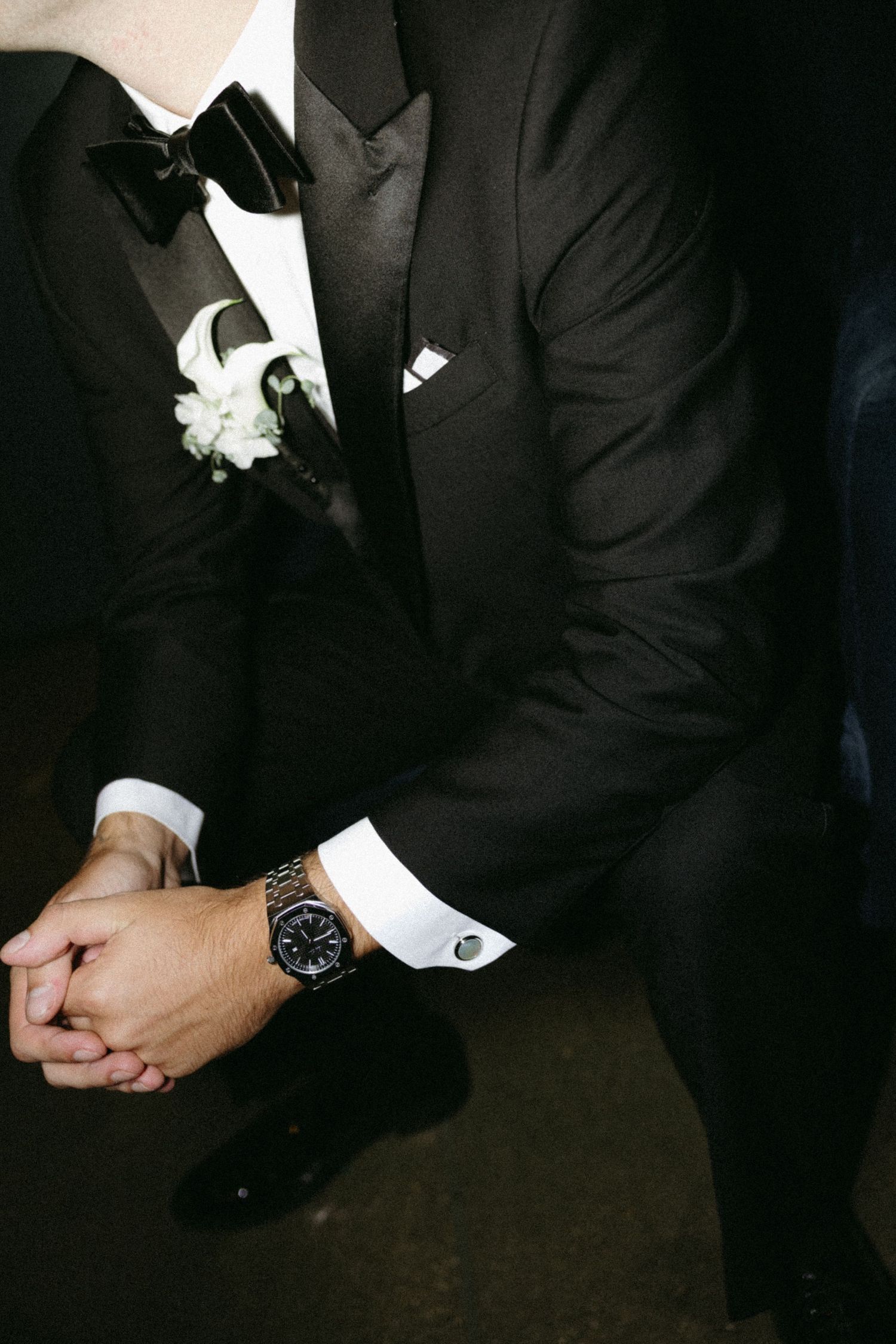 Two grooms in black tuxedos with white boutonnieres and cufflinks sit together, hands clasped, at a formal wedding.