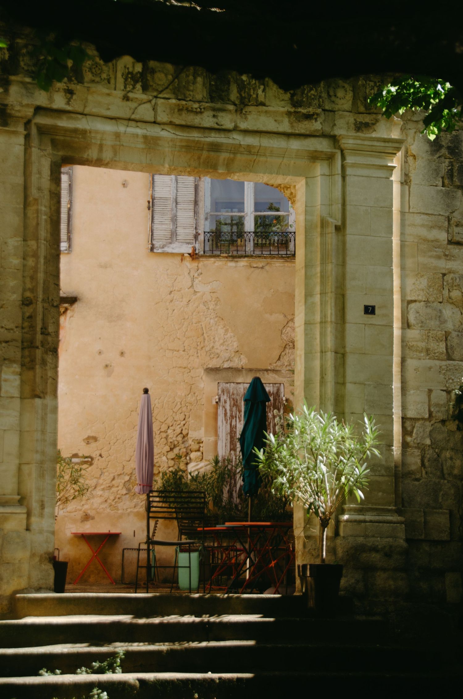 Ancient stone archway frames a weathered doorway with peeling paint and potted plants in a Mediterranean setting.