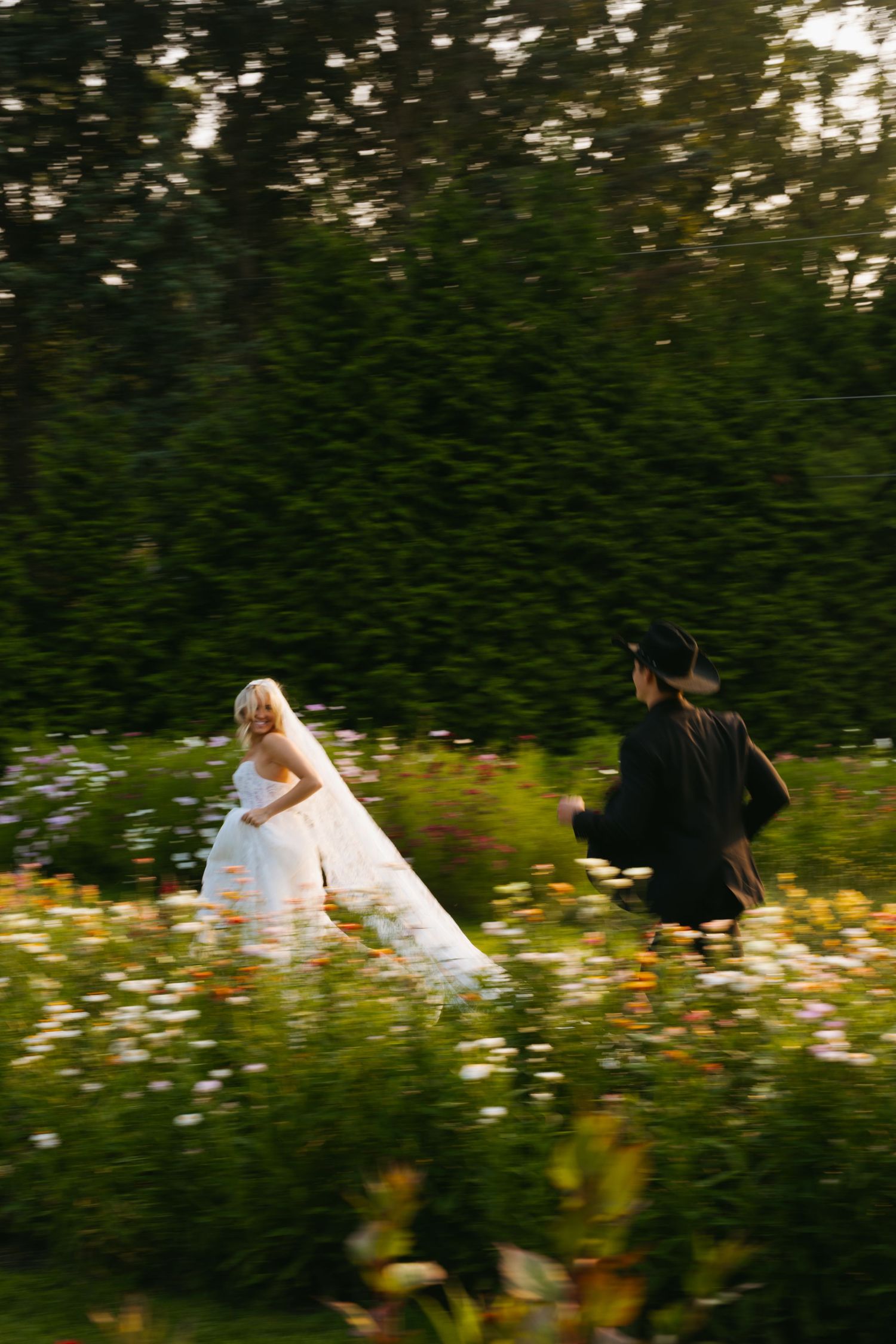 Two people in ethereal attire run through a wildflower meadow at golden hour with evergreen trees in background.