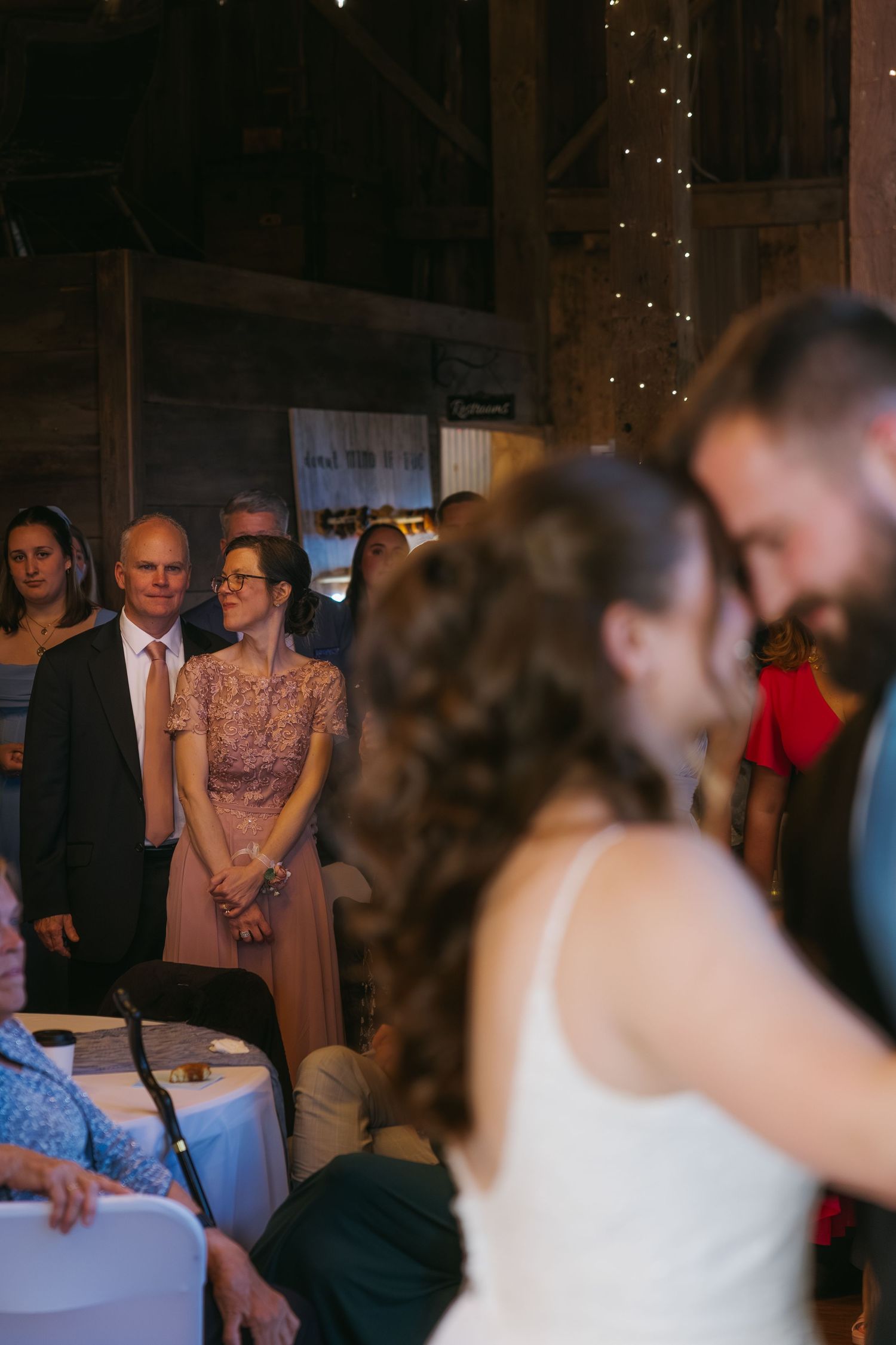 Guests watch bride and groom share first dance at rustic barn wedding reception with string lights.