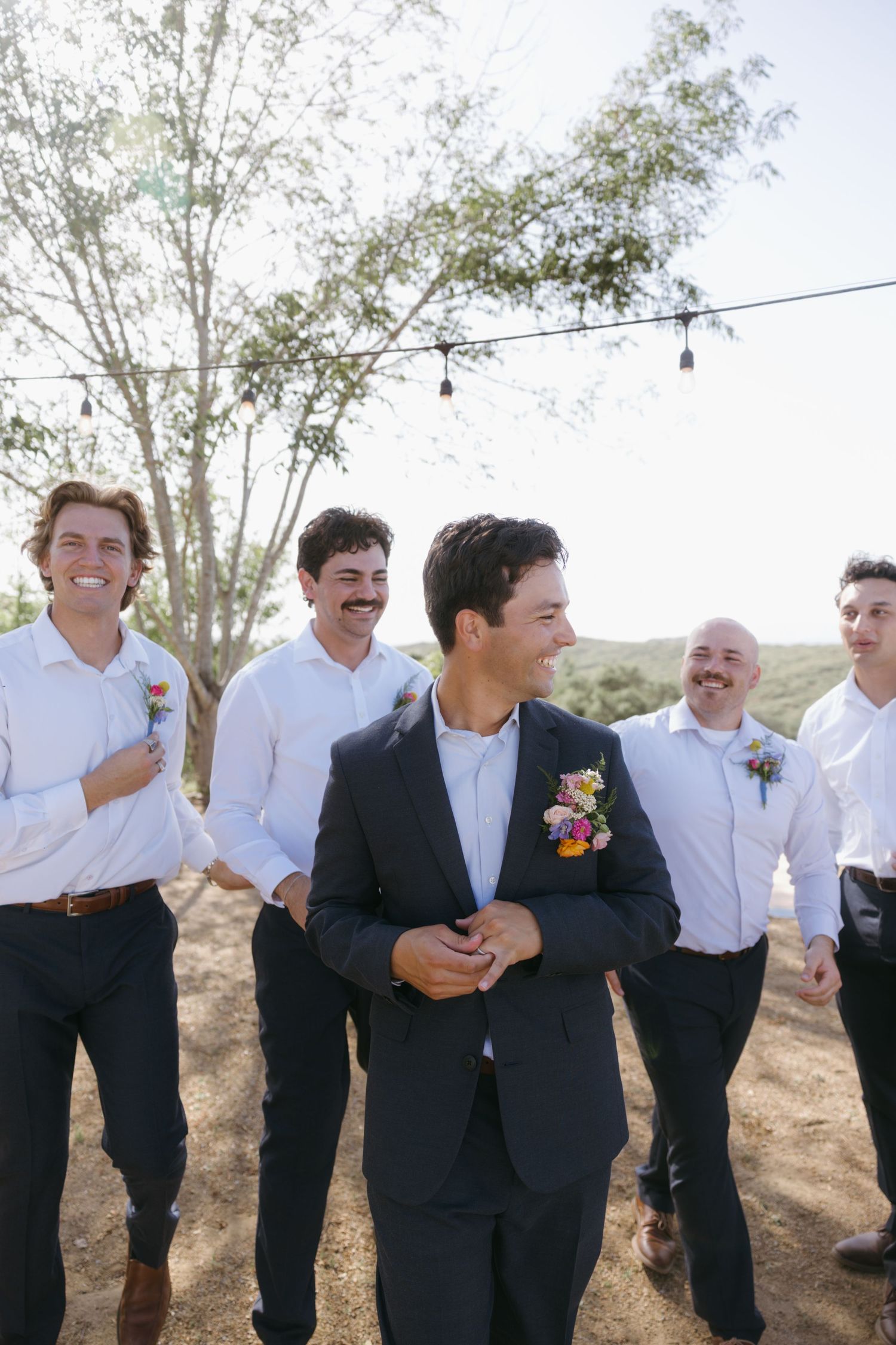 Groom in navy suit with floral boutonniere laughing with groomsmen in white shirts outdoors.