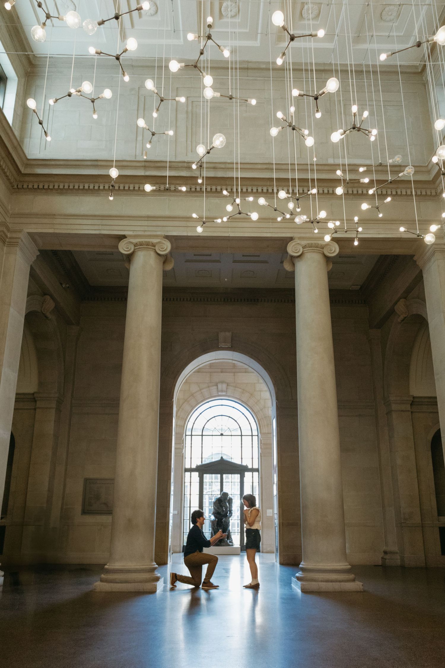 Modern chandelier installation hangs from ornate architecture ceiling above grand arched entryway with columns.