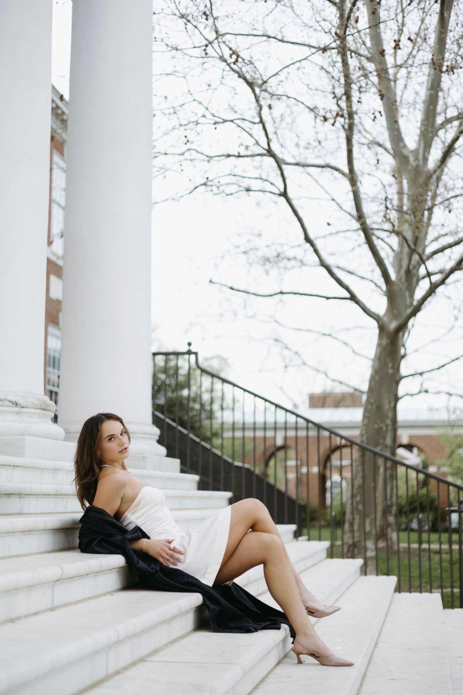 Woman in white dress and black cardigan relaxes on elegant stone steps near a bare tree in spring.