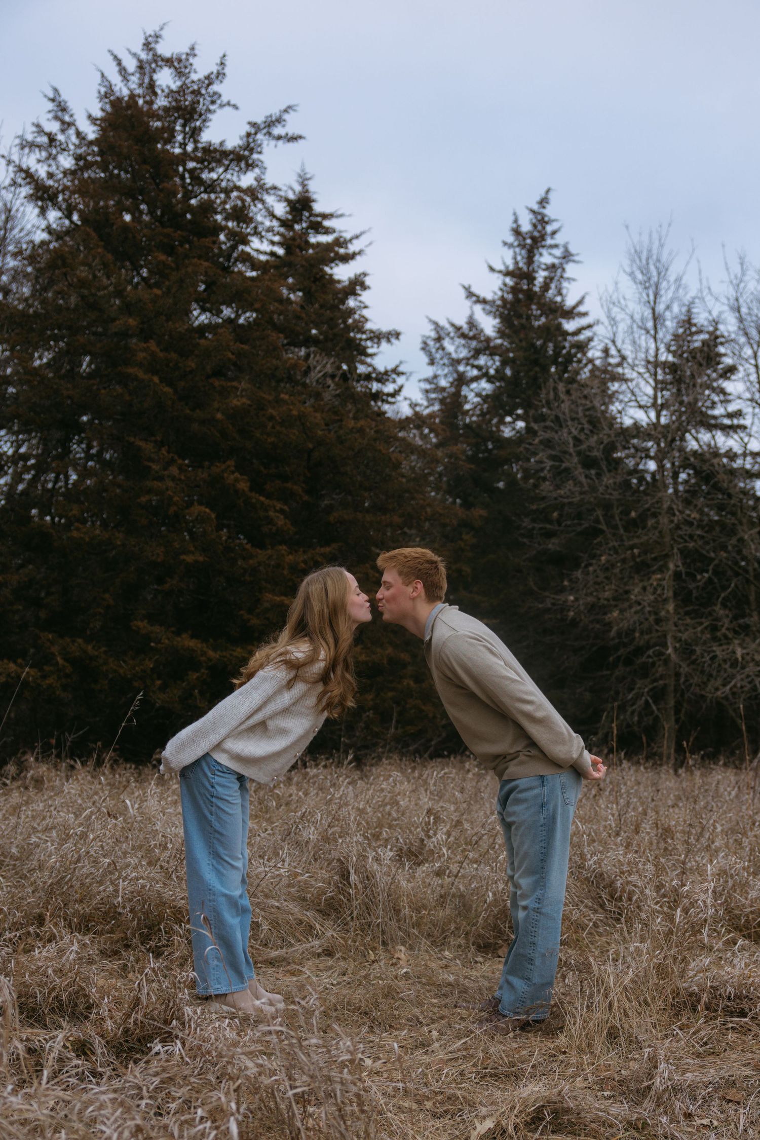 A couple in denim leans in for a kiss while standing in a brown grassy field with evergreen trees in the background.