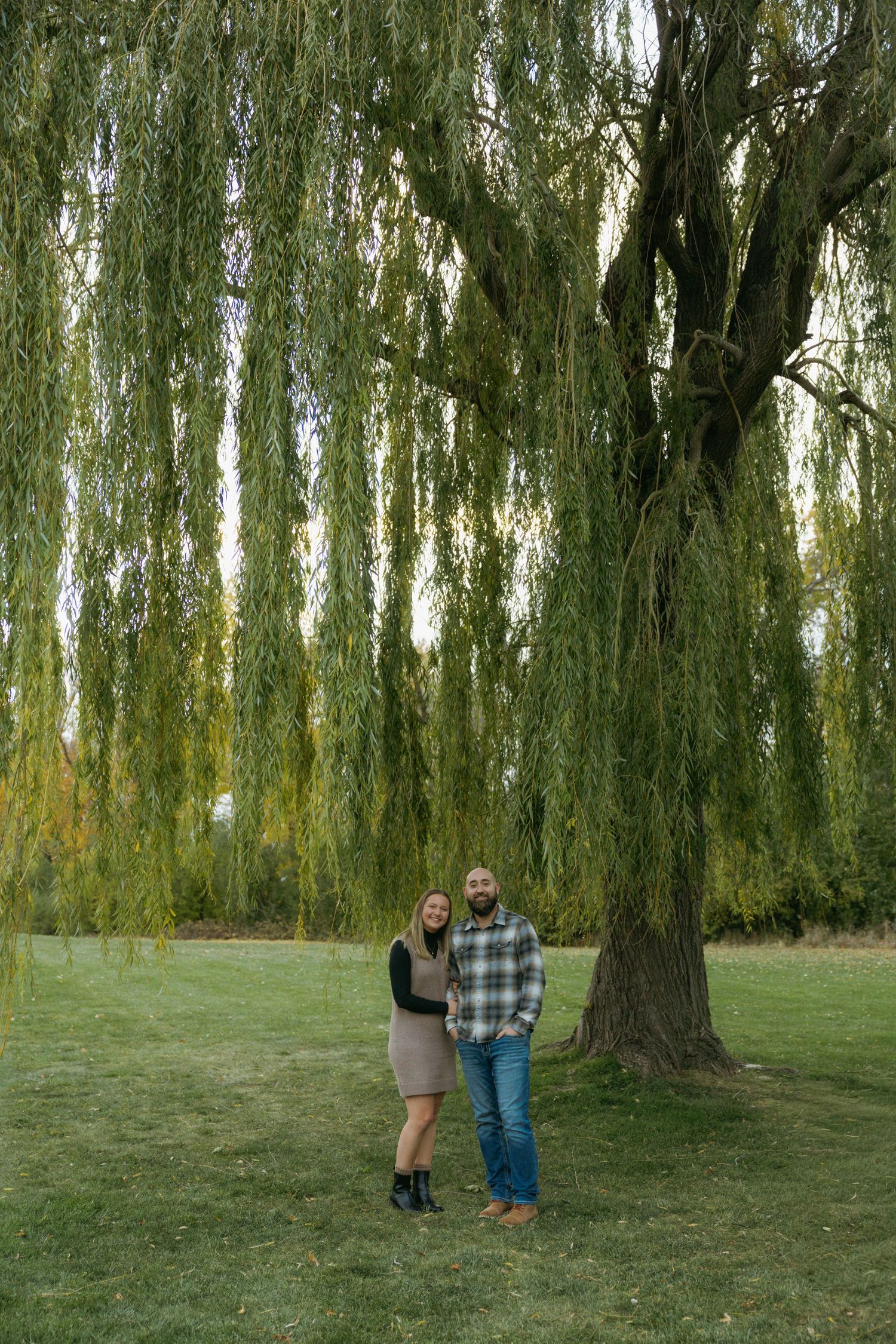 A couple stands beneath cascading willow trees in a serene park setting during a photo session.
