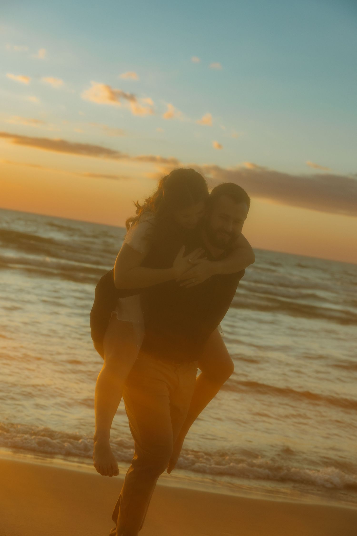 Silhouetted couple embracing on beach at sunset with golden light and ocean waves in background.