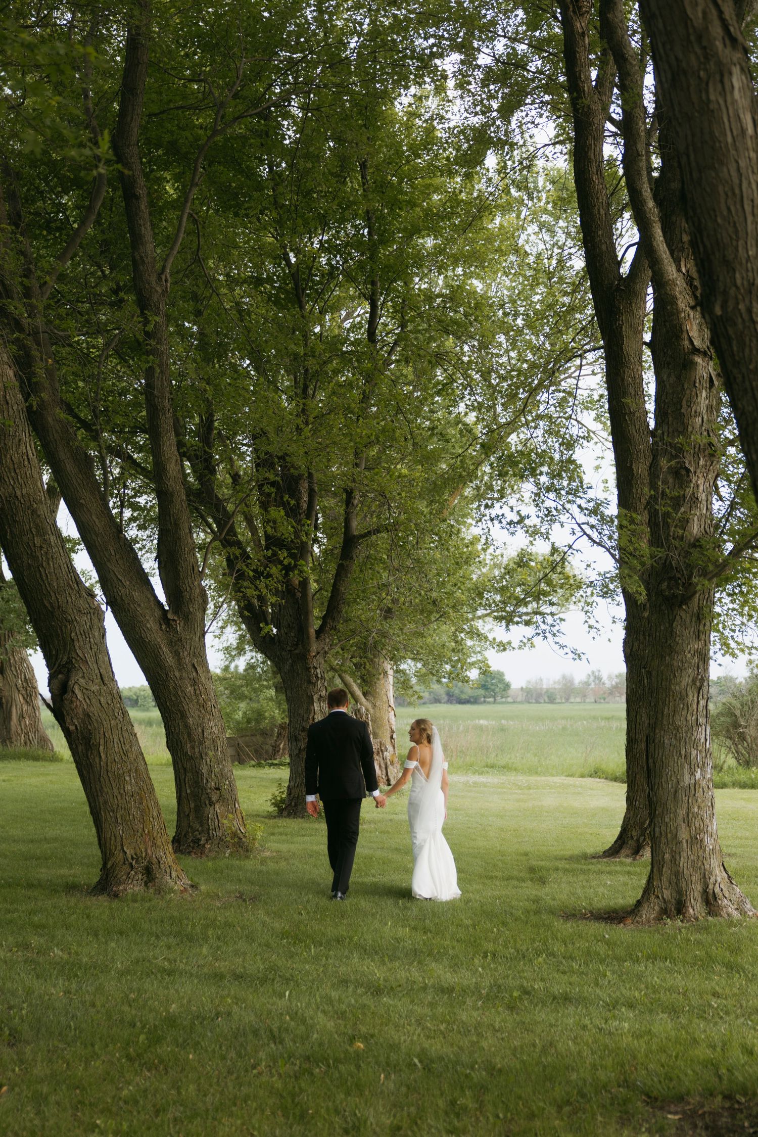 A couple walks through a scenic grove of mature trees on grass in an outdoor setting.