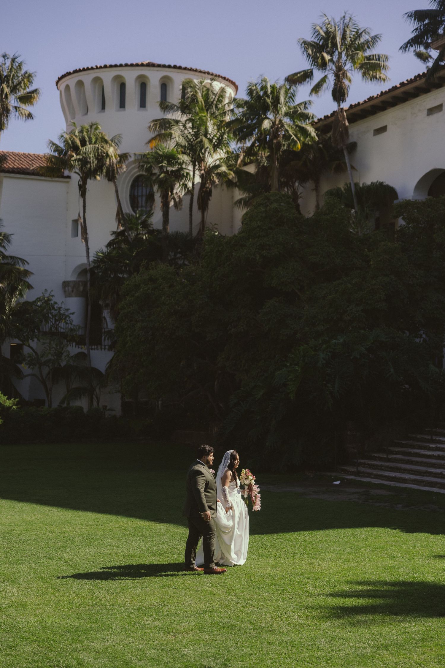 Bride and groom share a romantic moment on lush green lawn outside a white Spanish Colonial building with palm trees.