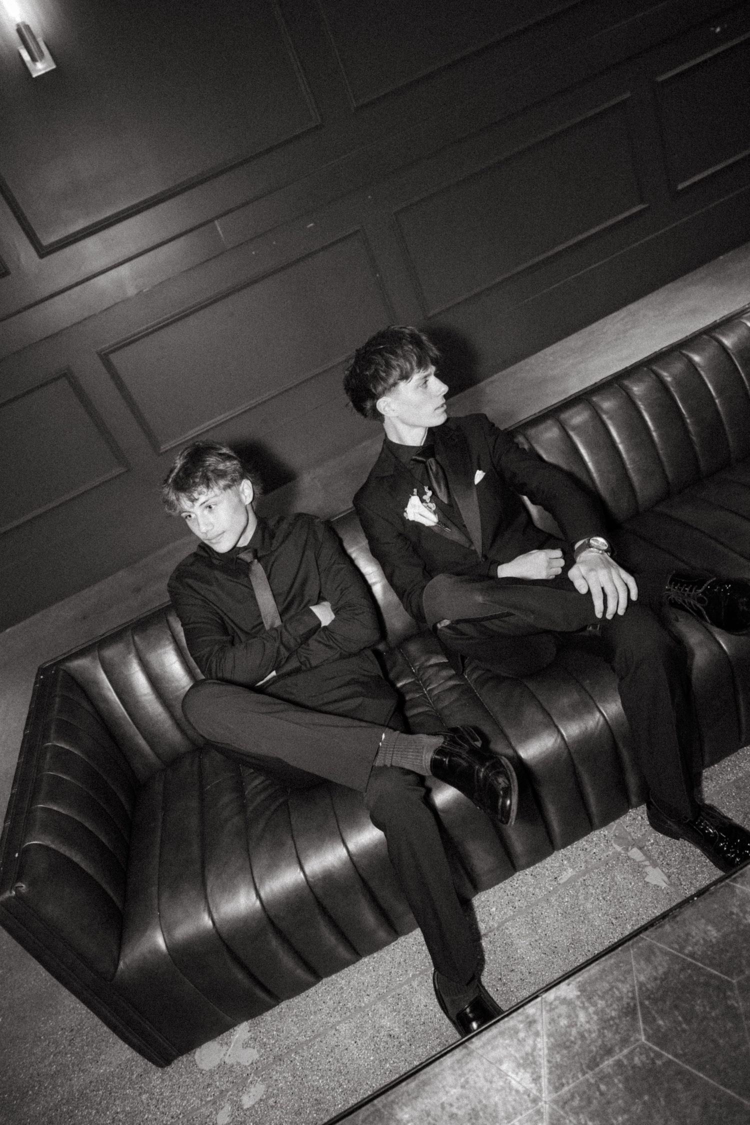 Two young men in formal suits sit on a leather sofa in a dimly lit room, black and white photo.