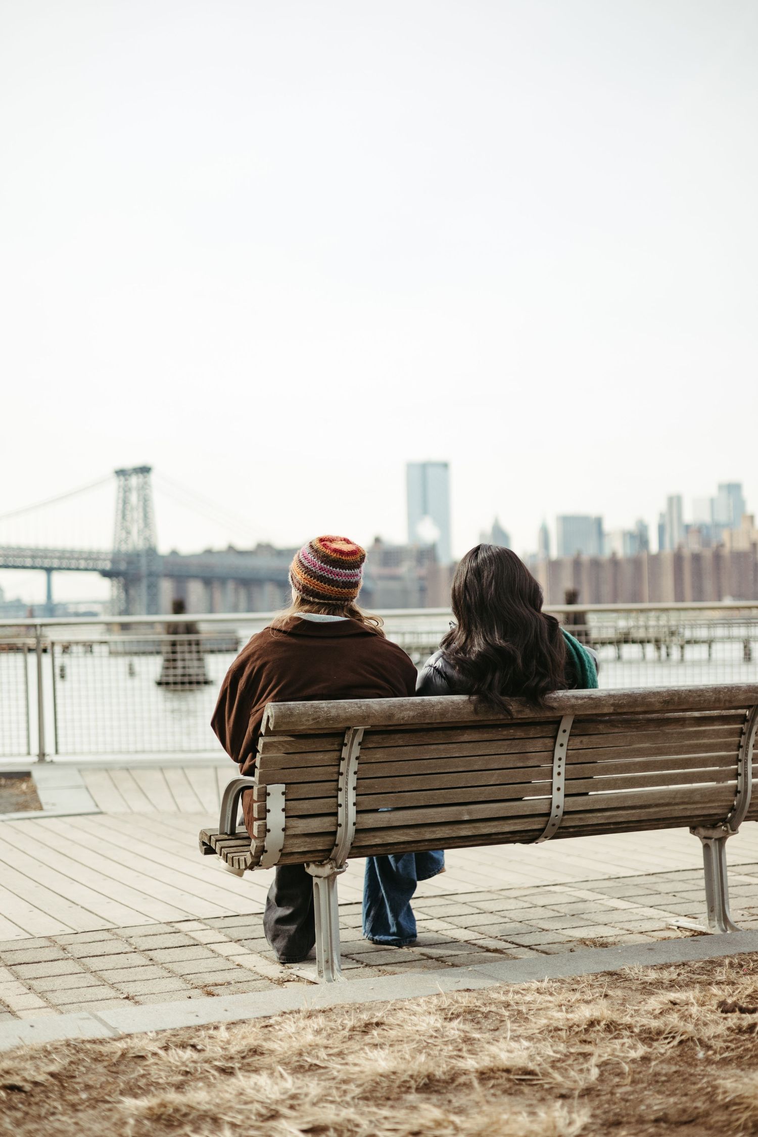 Two people sit on a wooden bench overlooking the Williamsburg Bridge and Manhattan skyline on a cloudy day.