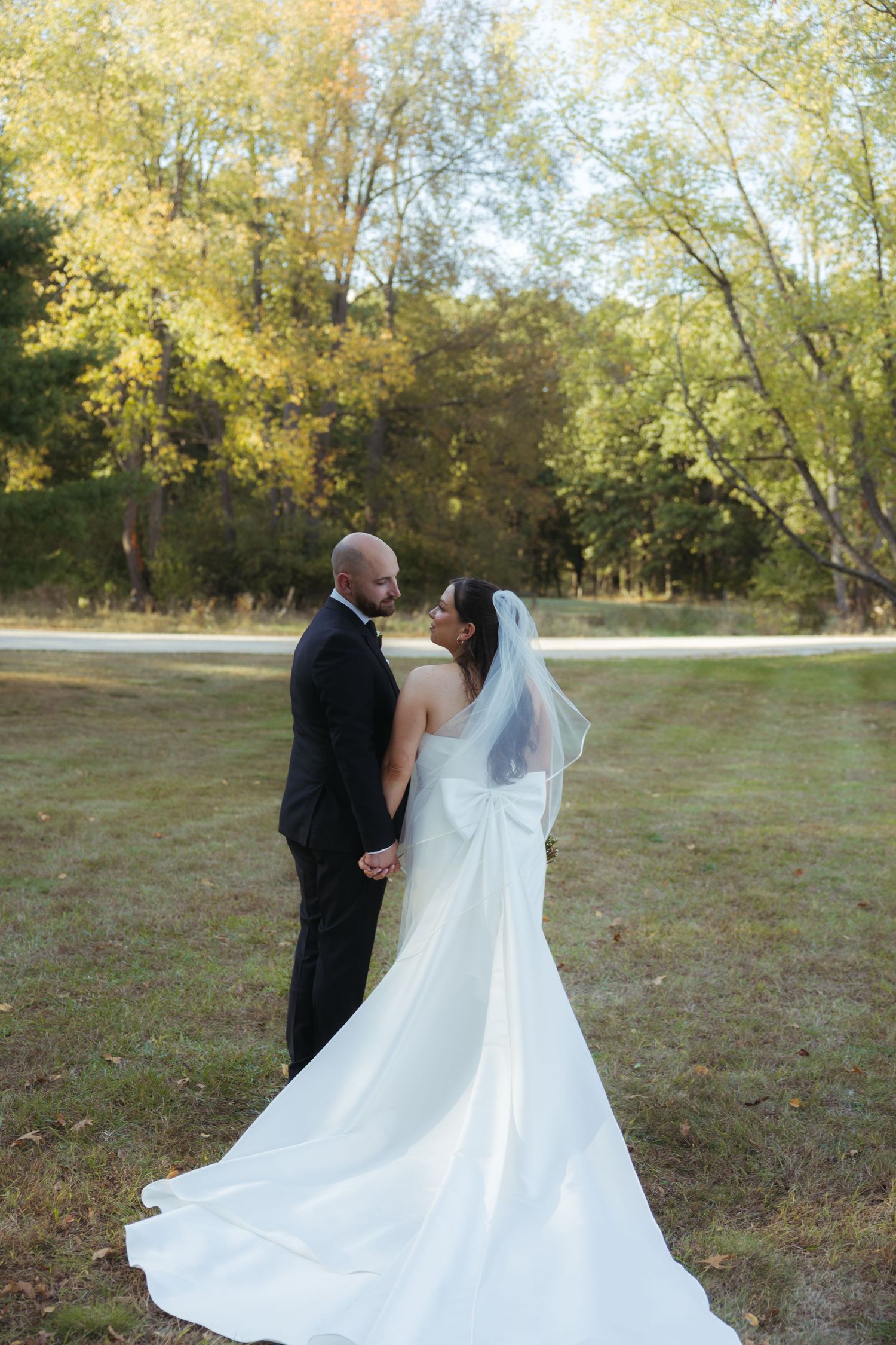 A couple in wedding attire stands together on an autumn day with golden trees in the background.