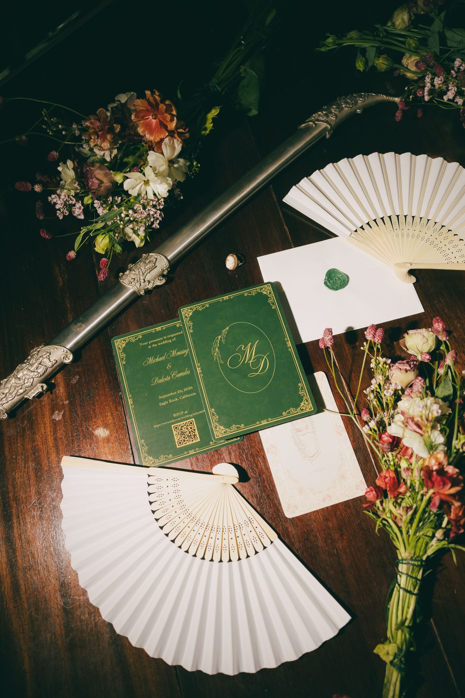 Elegant wedding stationery flat lay with green invitation, white fans, wax seal, and floral arrangements on wooden table.