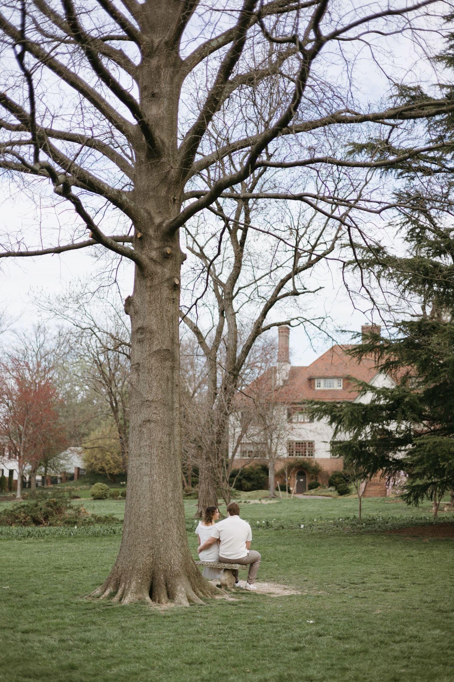 Two people in white sit together on a bench under a large bare tree on a green lawn with a house visible in the background.