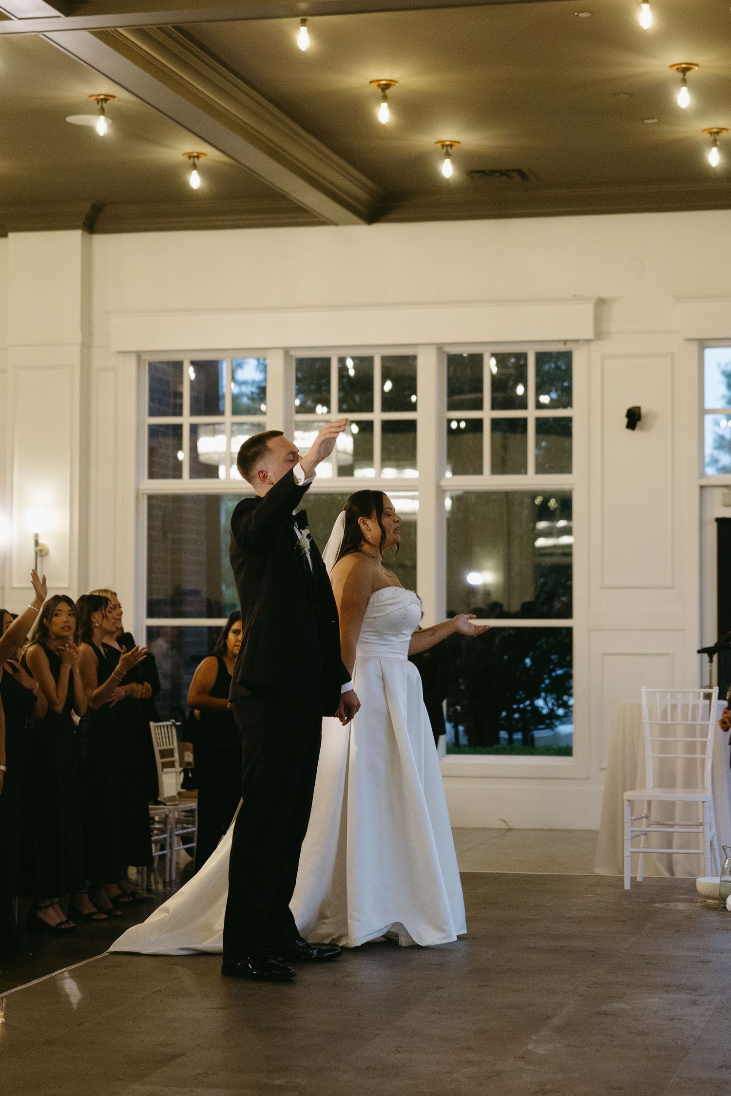 Bride and groom share their first dance in an elegant white reception hall with string lights and guests watching.