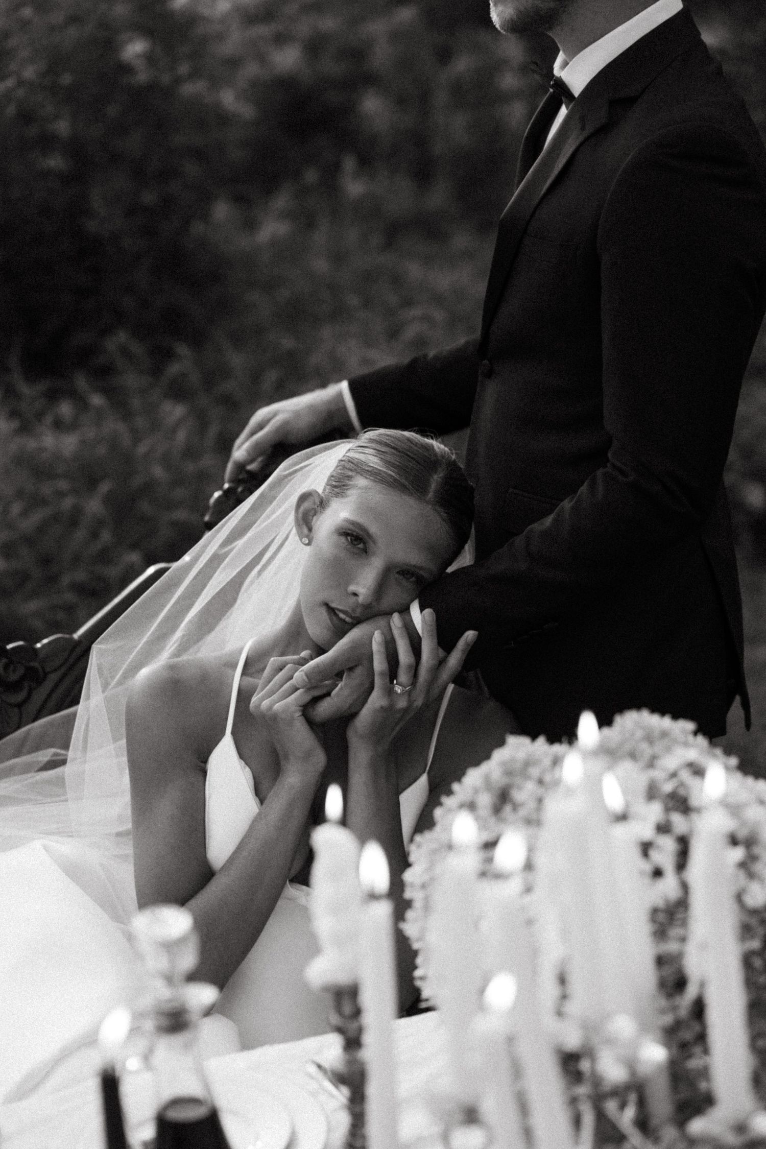 Black and white wedding photo with a bride in white dress resting her head near candles while groom stands behind her.
