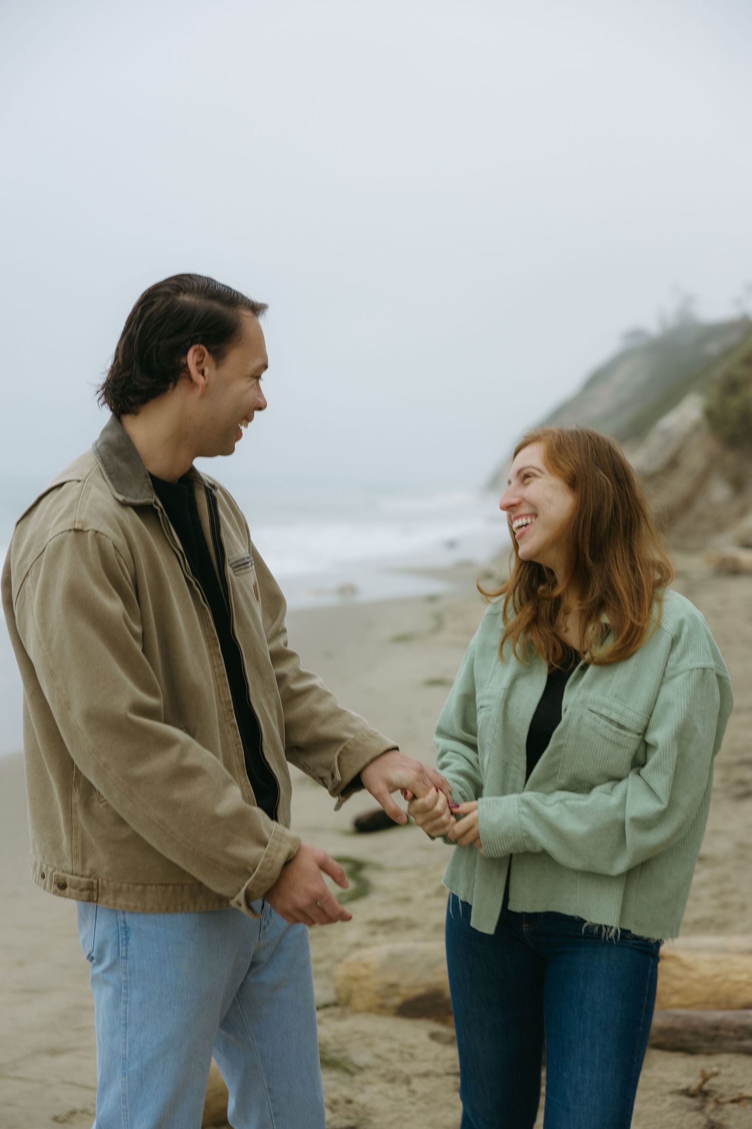 A couple shares a playful moment while walking along a misty beach in casual fall attire.