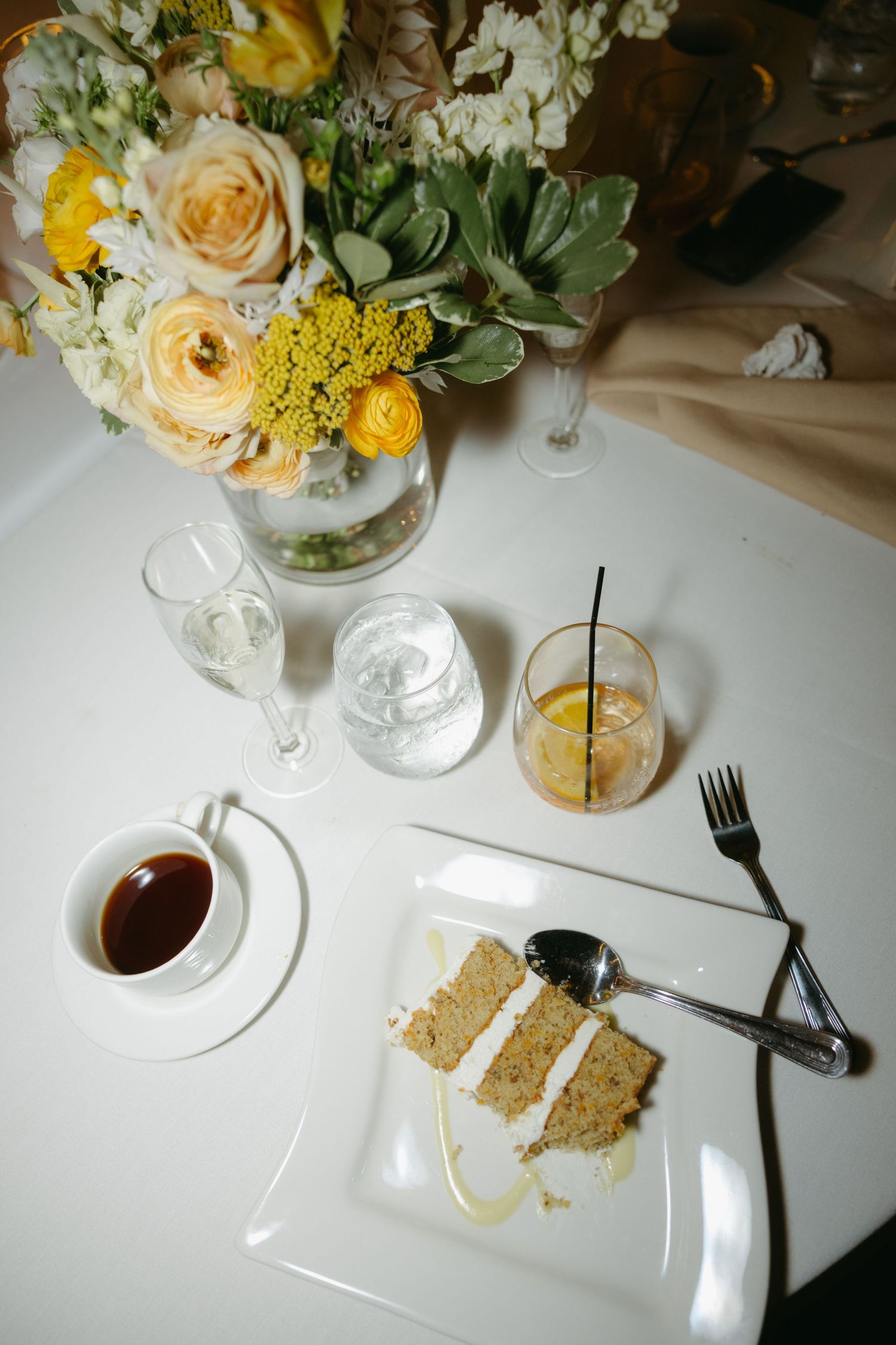 Elegant wedding reception table with floral centerpiece, slice of layered cake, coffee cup, and cocktail on white tablecloth.