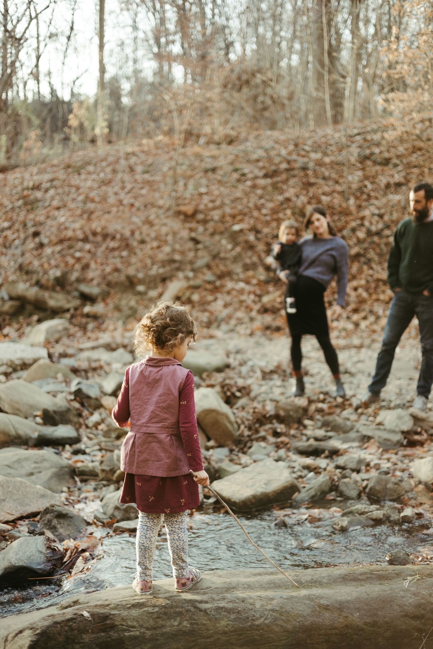 Child exploring a rocky creek bed while others watch from behind in a wooded setting.