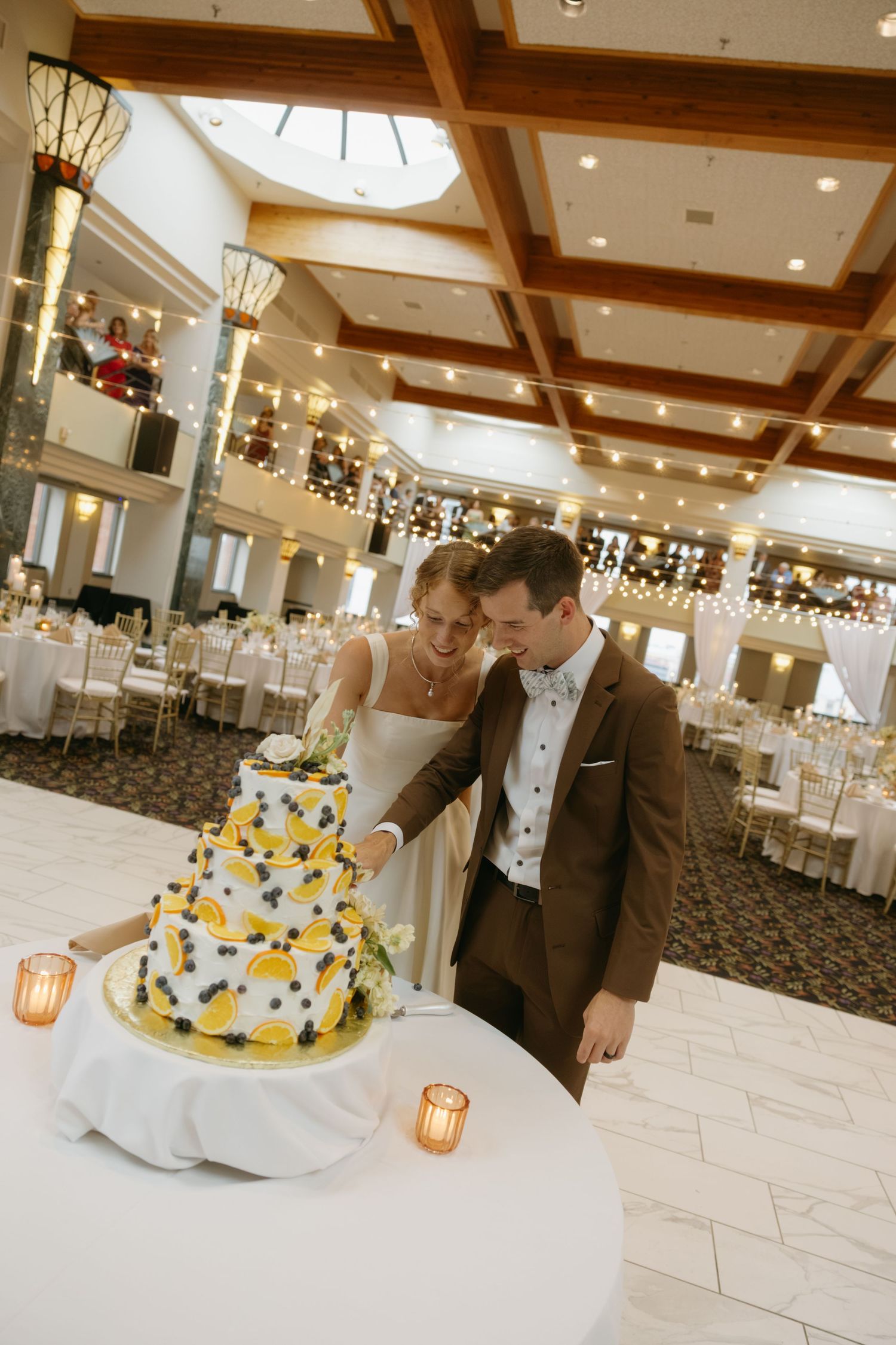 Bride and groom cutting a three-tier yellow and black wedding cake in an elegant reception hall with string lights.