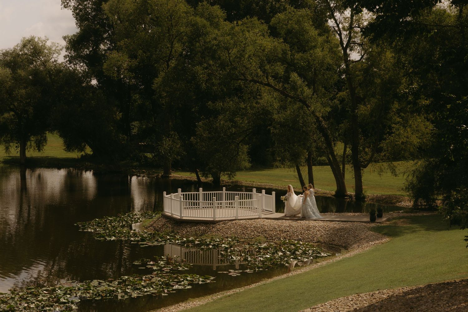 Bride in flowing white gown and veil stands on ornate bridge over lily pad pond surrounded by lush green trees.