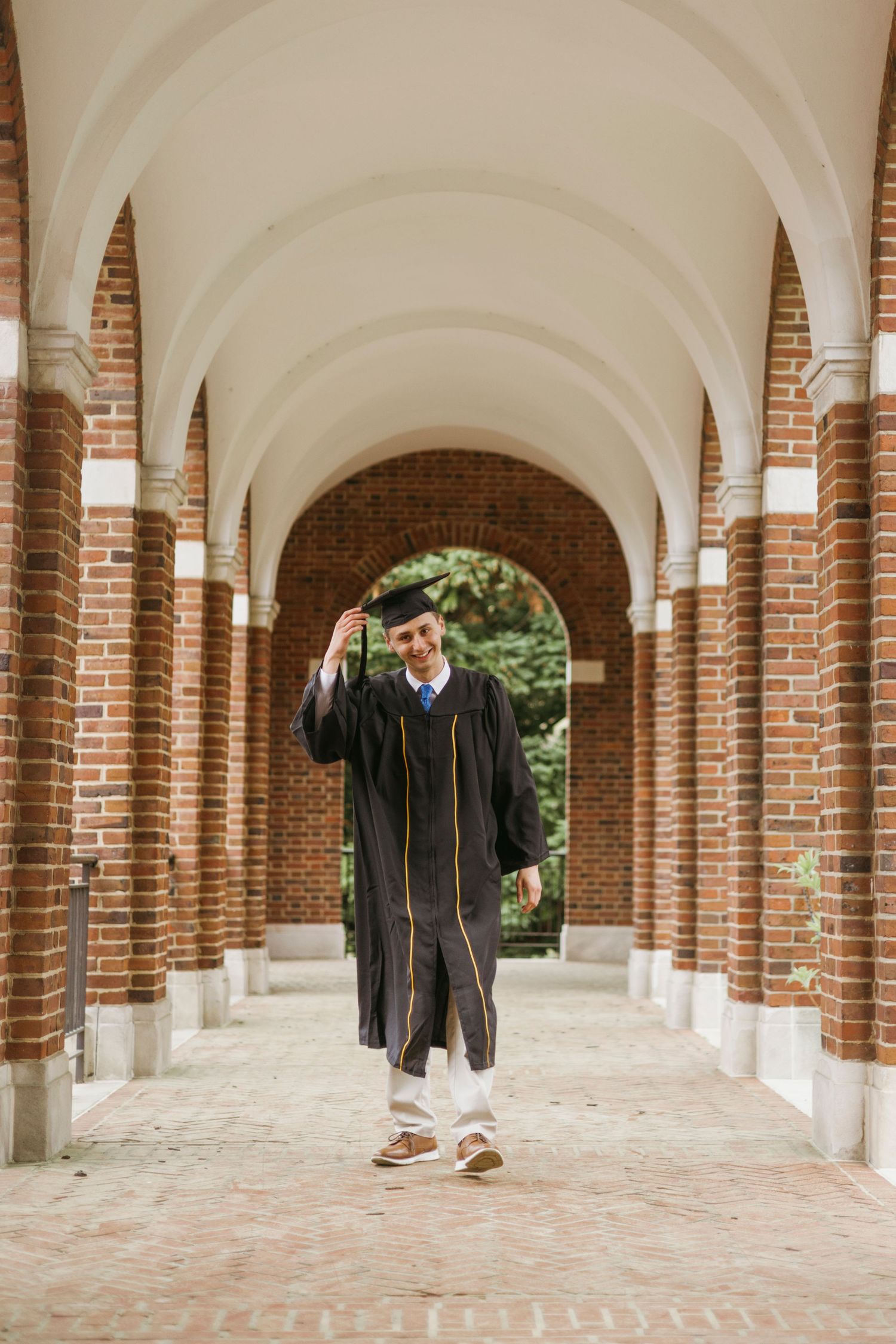 Graduate student wearing black cap and gown stands in a brick archway corridor during graduation celebration.