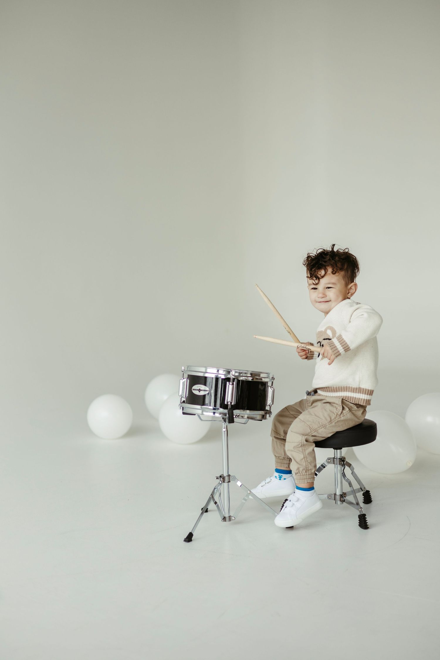 Young drummer in white sweater and khaki pants playing on snare drum in minimalist white studio setting with balloons.