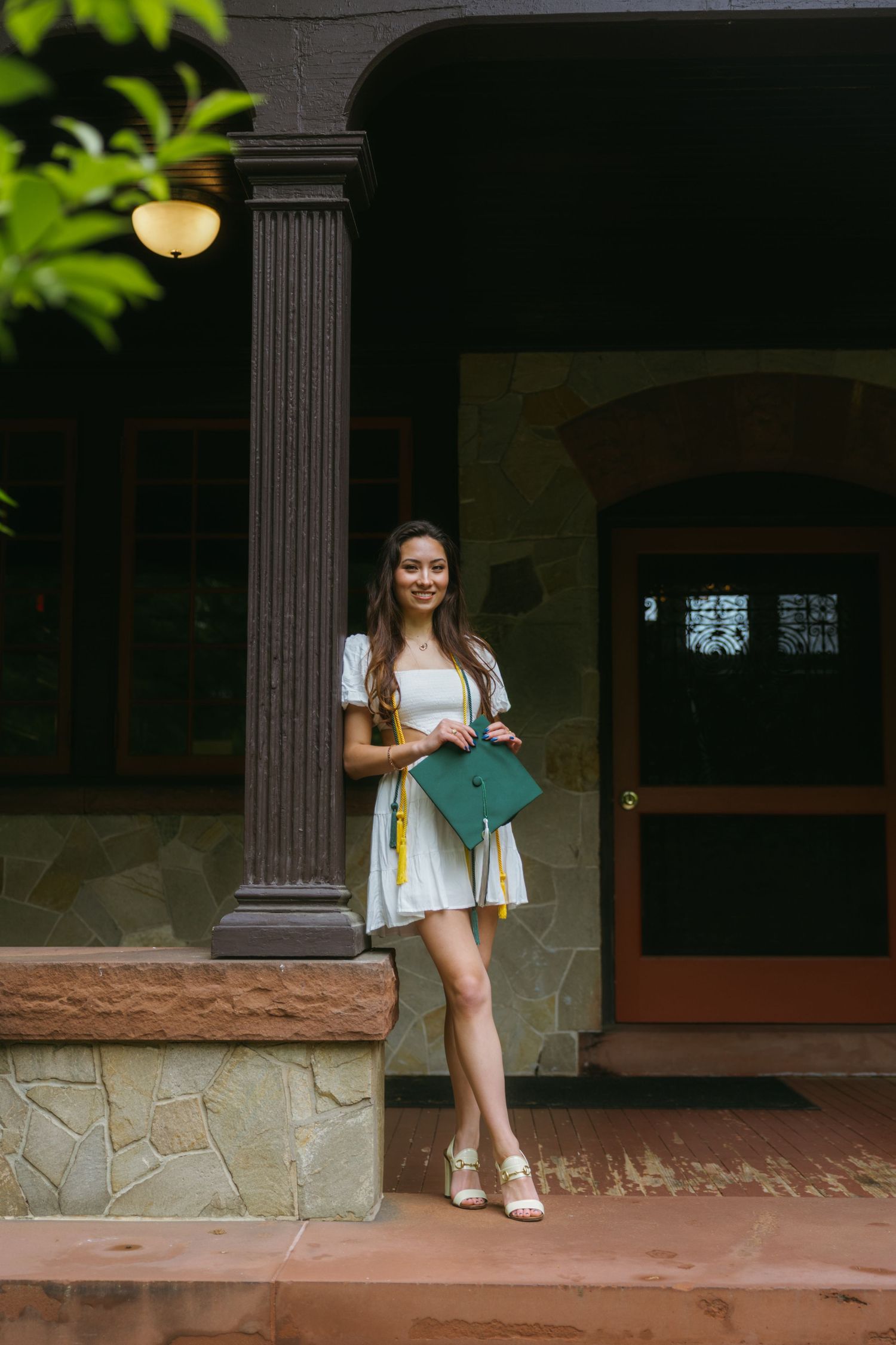 Young woman in white dress holding green graduation cap on stone porch, celebrating academic achievement.