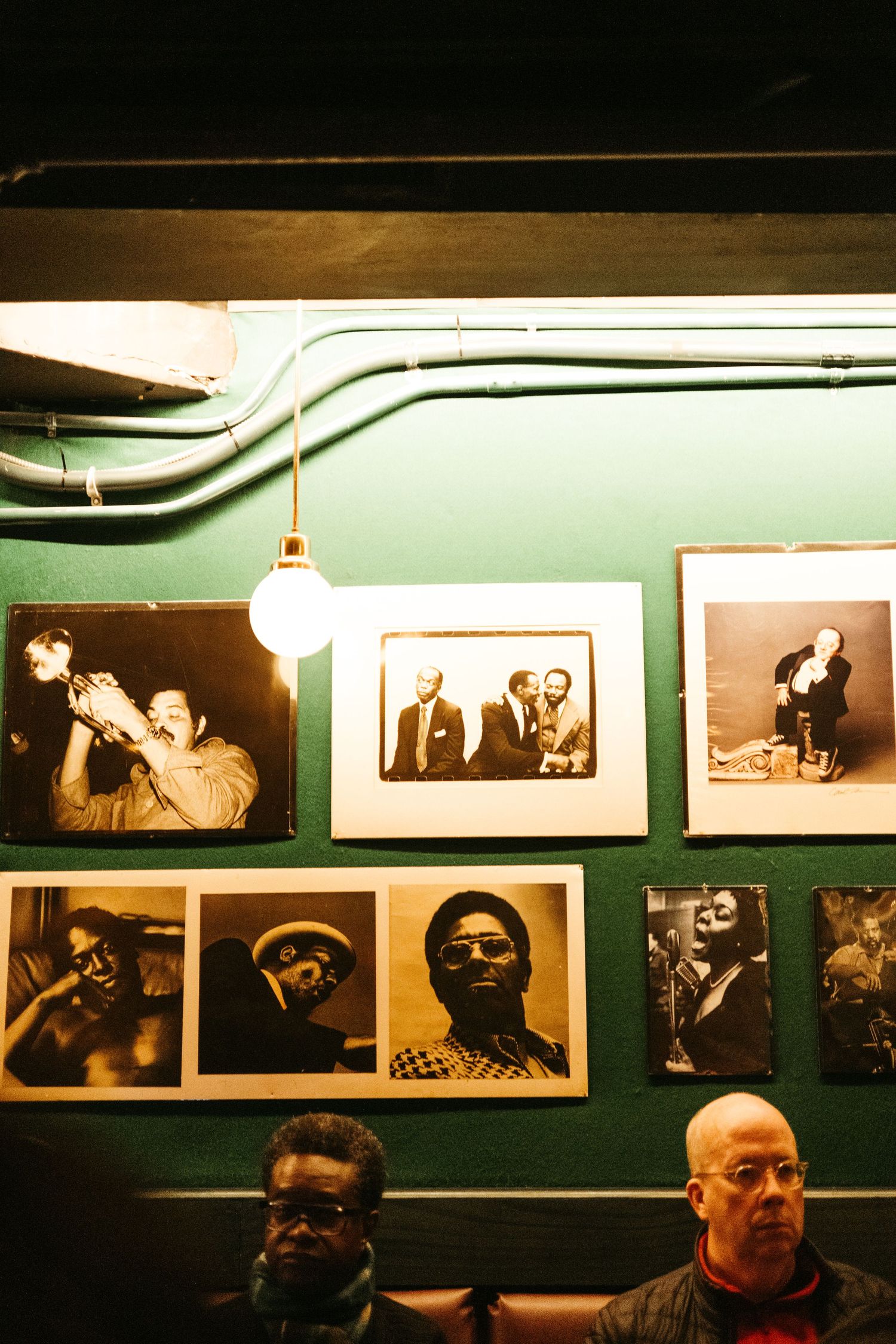 A wall of vintage jazz and blues photographs displayed above seated patrons in what appears to be a music venue.