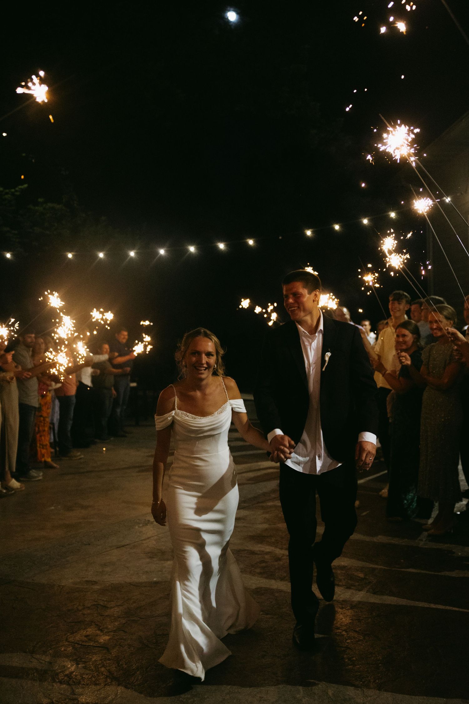 A couple walks beneath sparklers held by their wedding guests during an evening celebration outdoors under string lights.