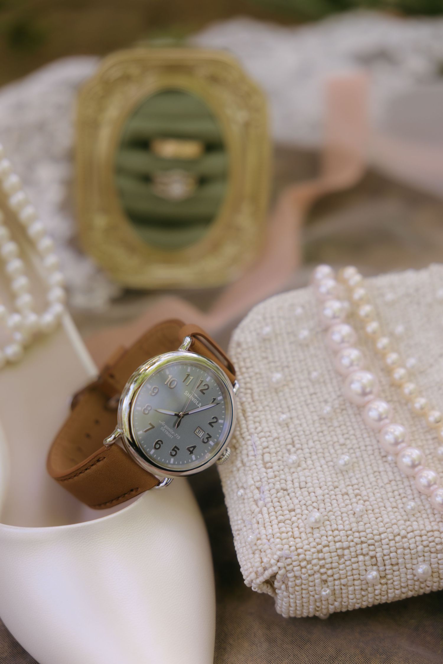 Elegant bridal accessories flat lay featuring a tan leather watch, pearl clutch, white heels, and gold ring box.