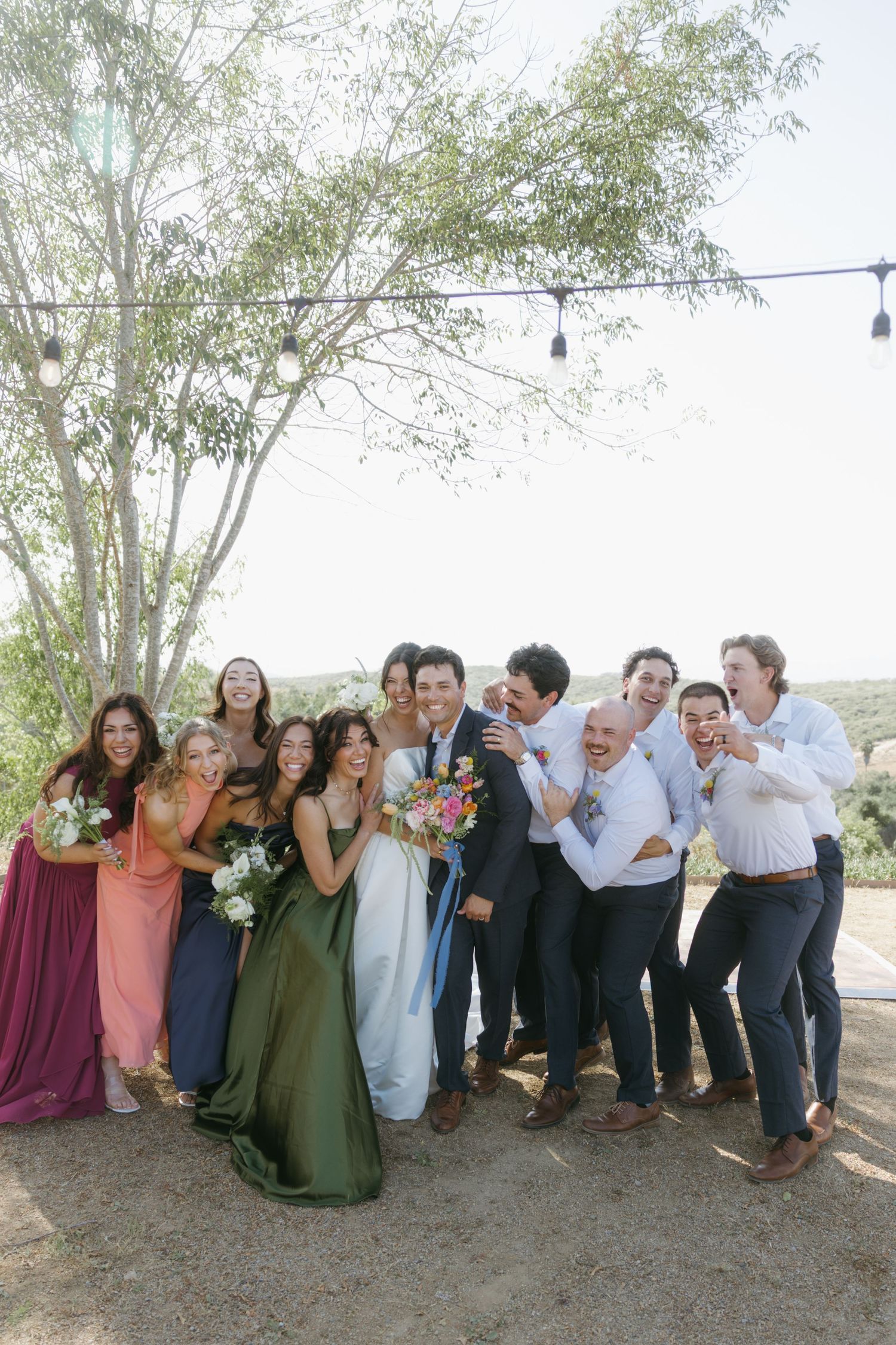 Wedding party posing outdoors, bride in white gown with colorful bridesmaids and groomsmen celebrating joyfully under string lights.