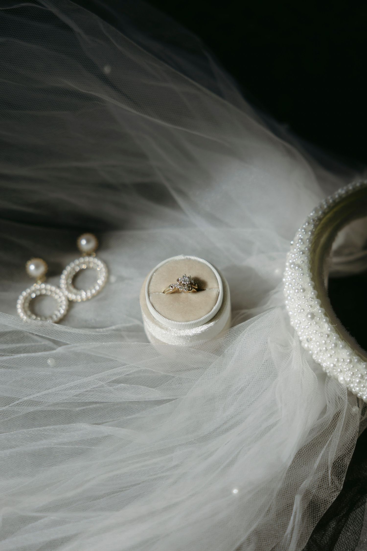 Wedding accessories including engagement ring in white box, pearl earrings and delicate veil on dark background.