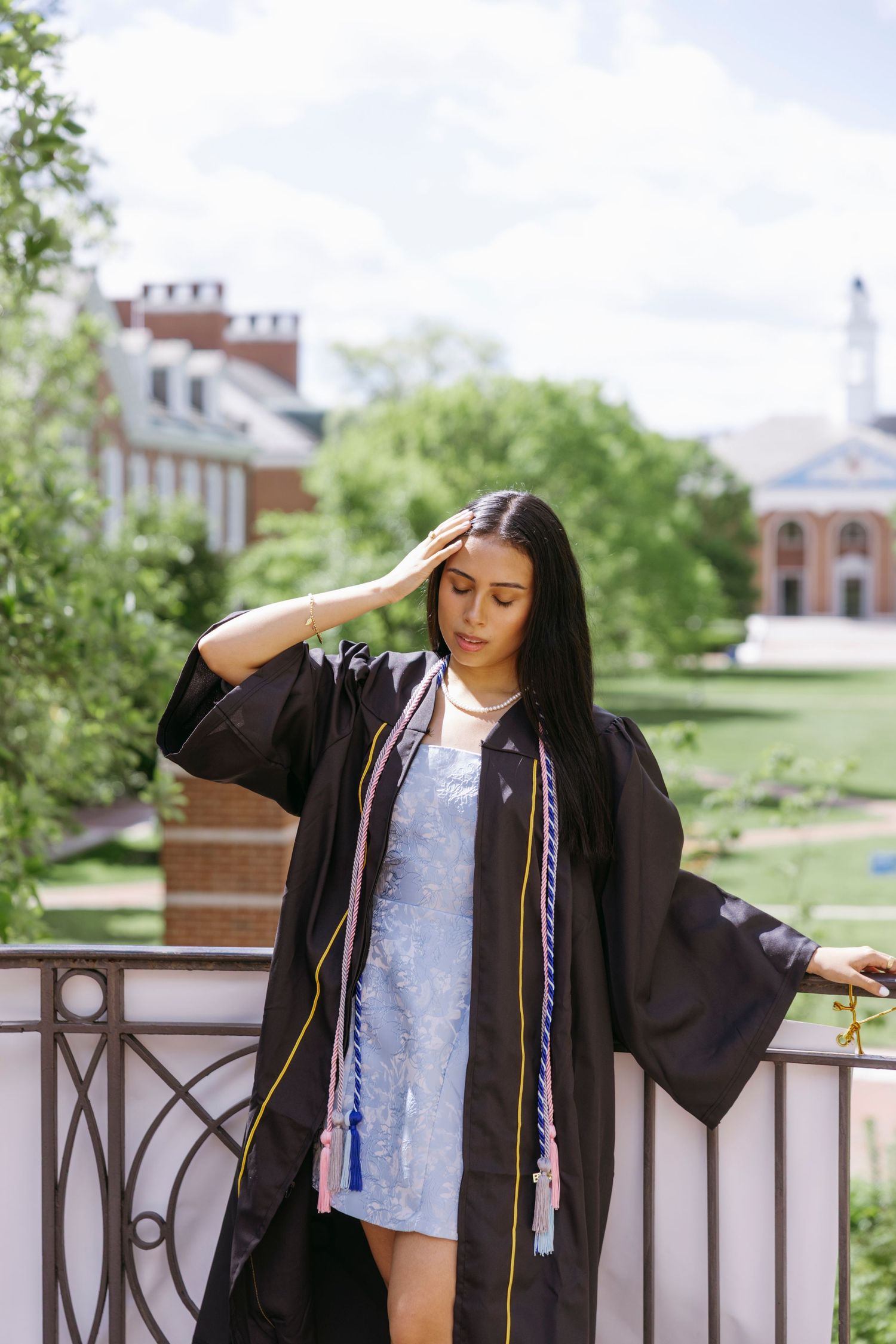 Graduate in cap and gown poses on campus balcony with brick buildings and white church steeple in background on sunny day.