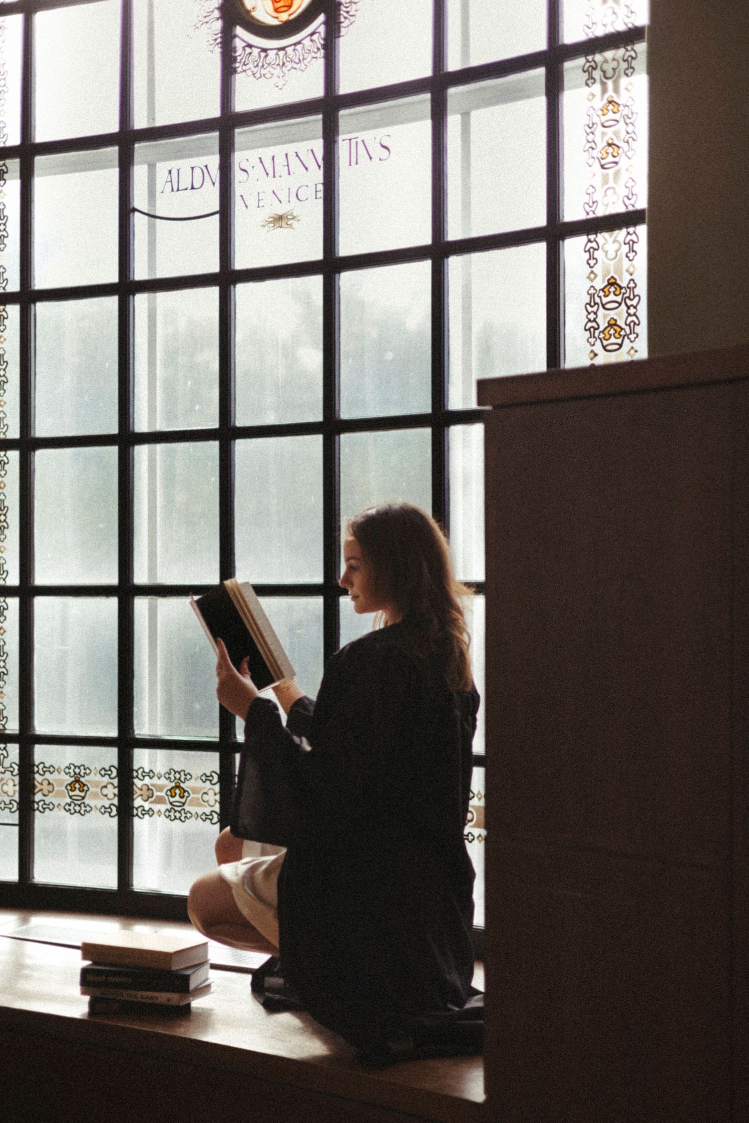 Silhouetted figure sits by window reading book in atmospheric moody room with vintage paned glass and decorative text above.