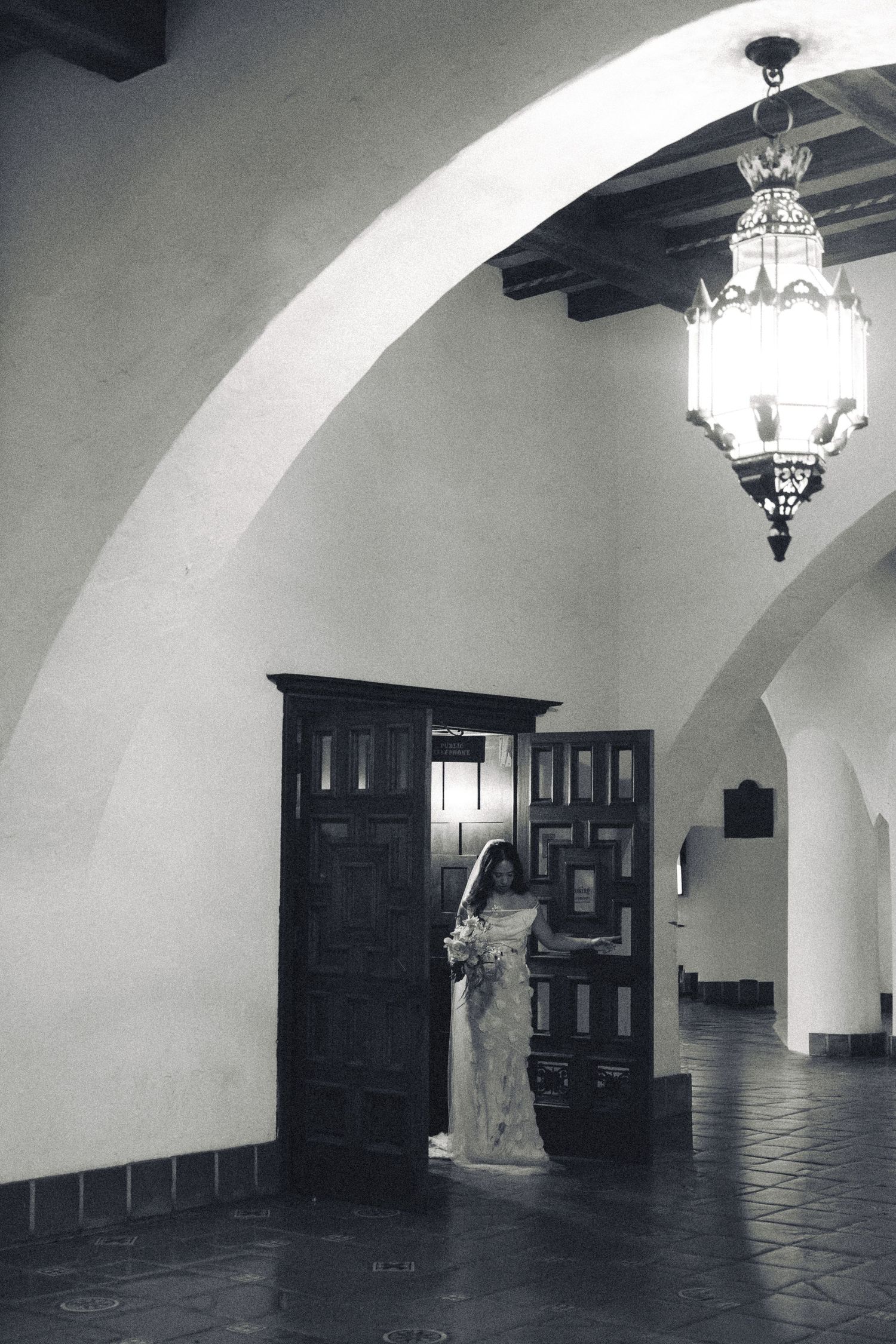 Black and white photo of a bride in a wedding dress holding flowers, standing by ornate wooden doors under an arched ceiling.