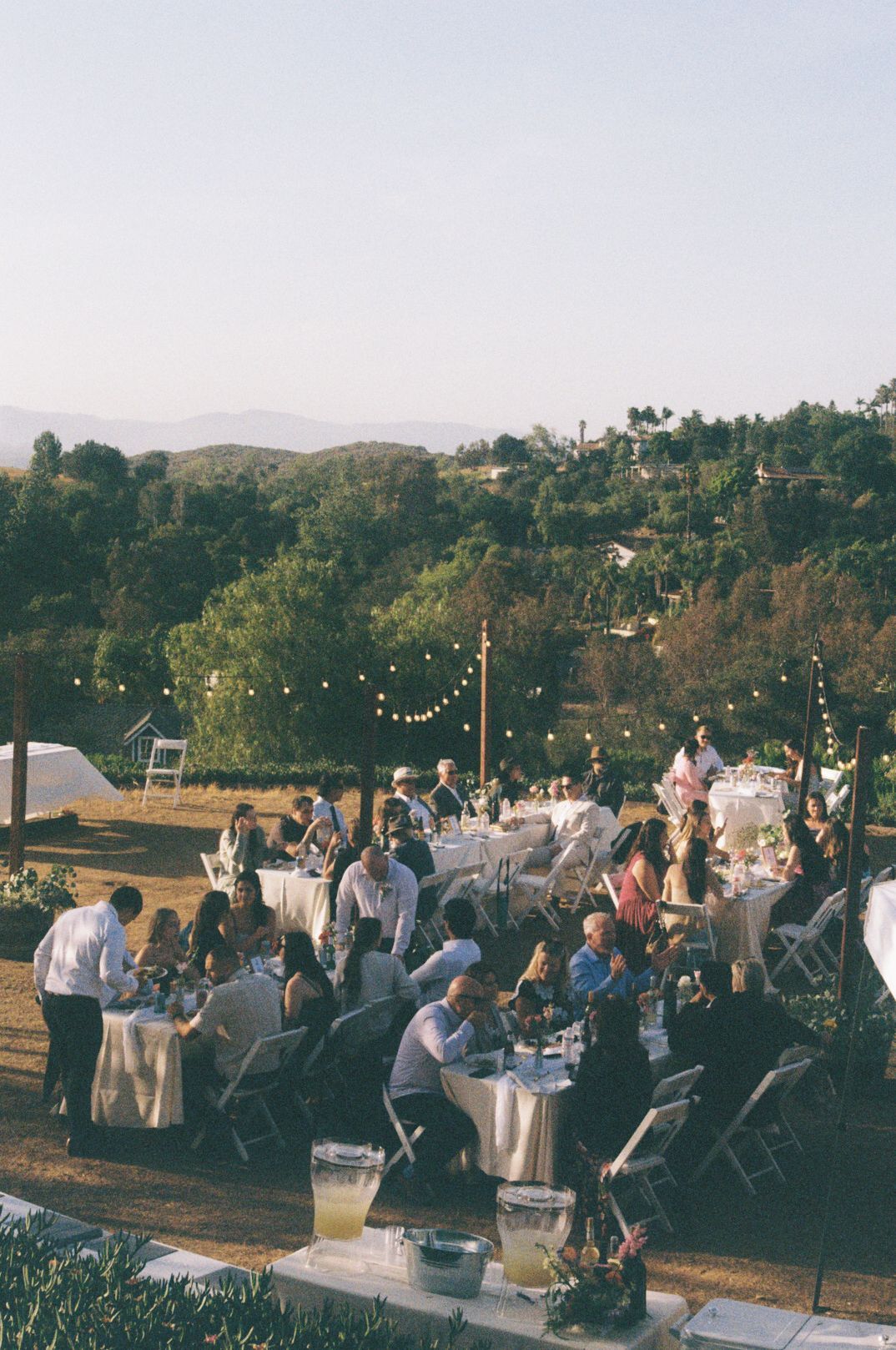 Outdoor wedding reception with string lights, guests dining at tables, surrounded by lush greenery and rolling hills at dusk.