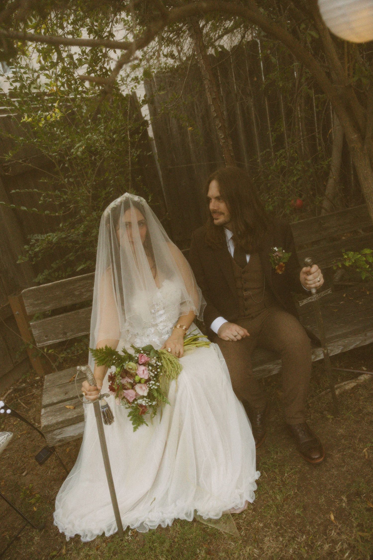 Bride in white gown and veil holds pink bouquet beside groom in brown suit on garden bench.