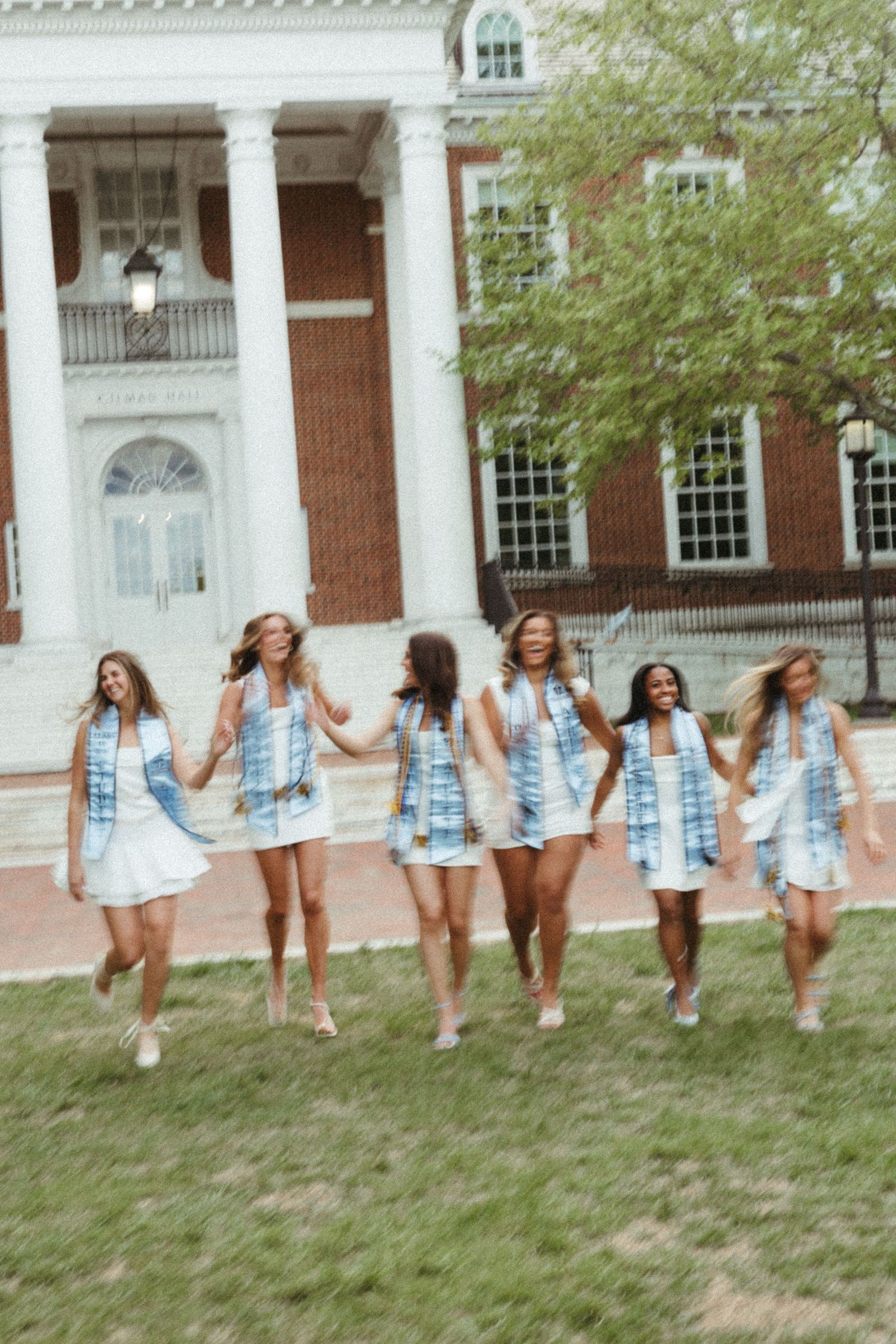 Six young women in white dresses and blue sashes walk together on a college campus lawn.