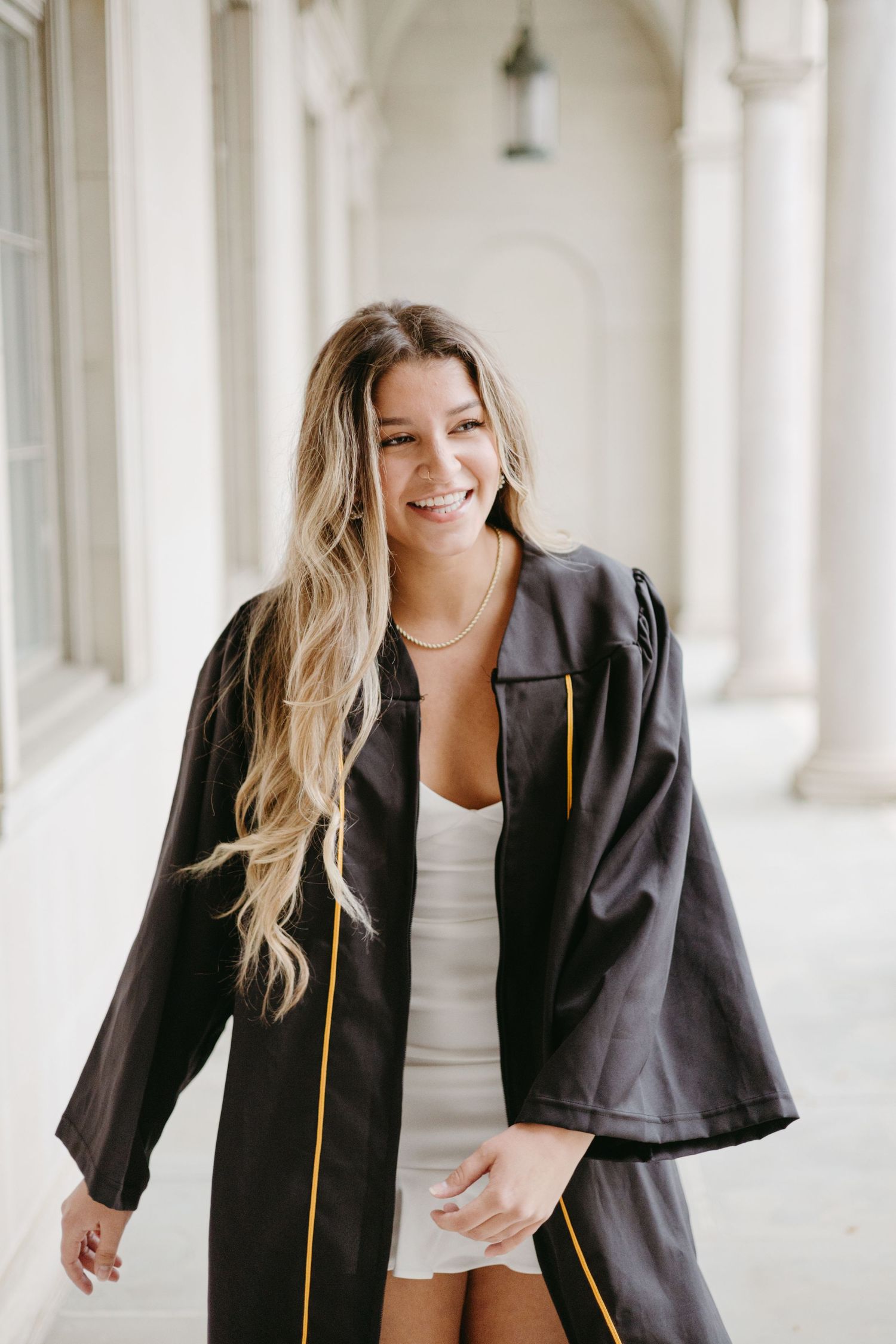 Graduate in black academic regalia poses in white columned hallway.
