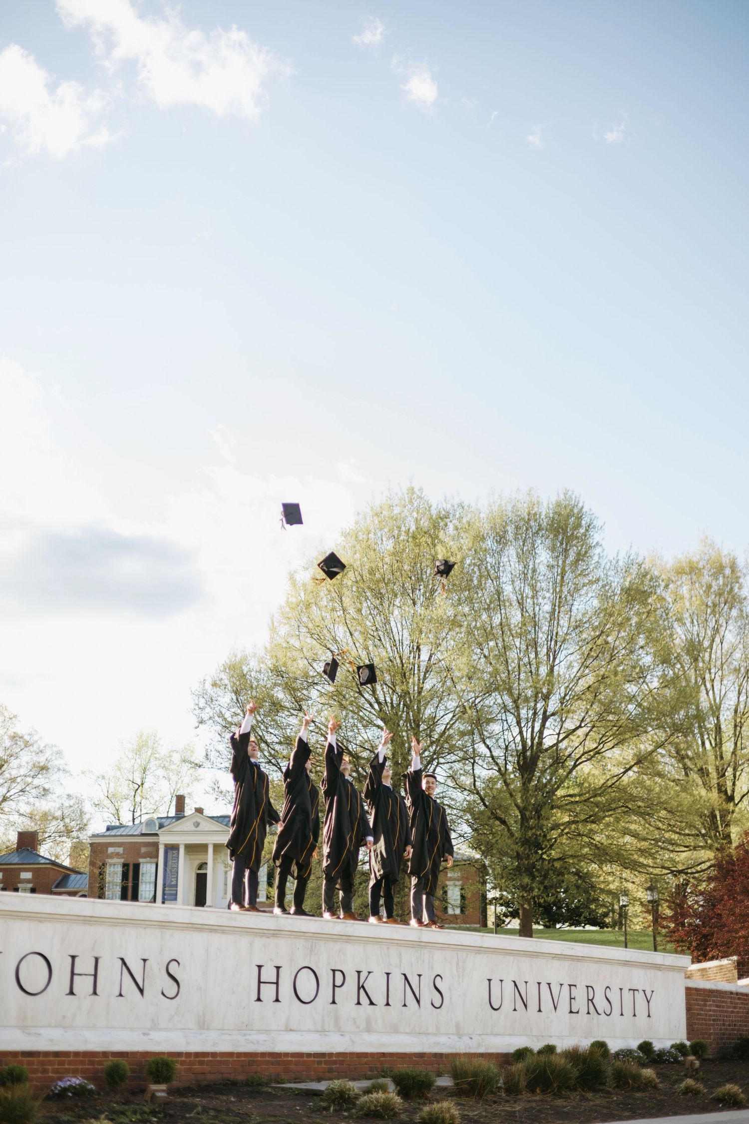 Graduates throw their graduation caps in the air by the Johns Hopkins University entrance sign on a sunny spring day.