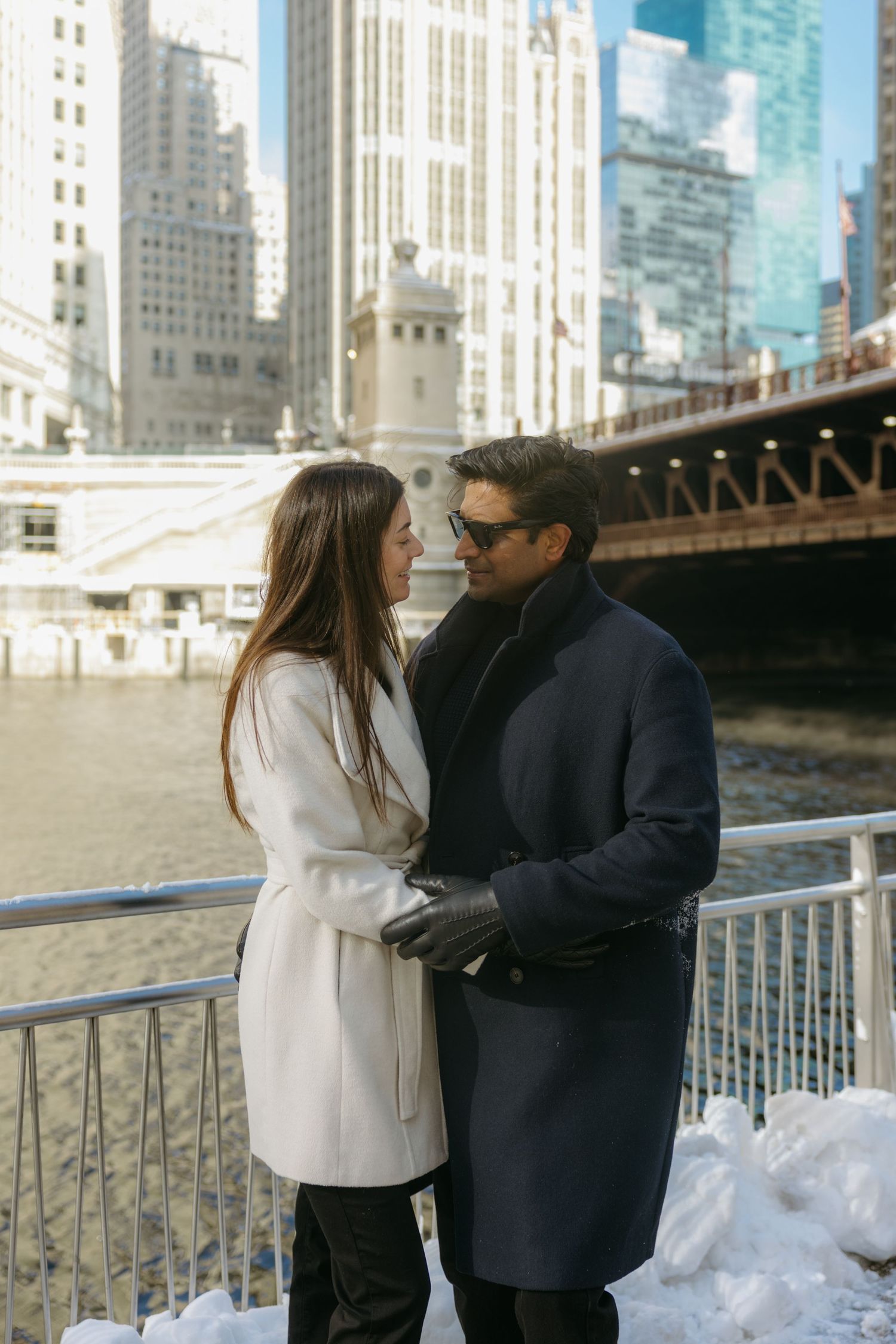 A couple shares a romantic moment along a snowy riverfront with city buildings in the background.