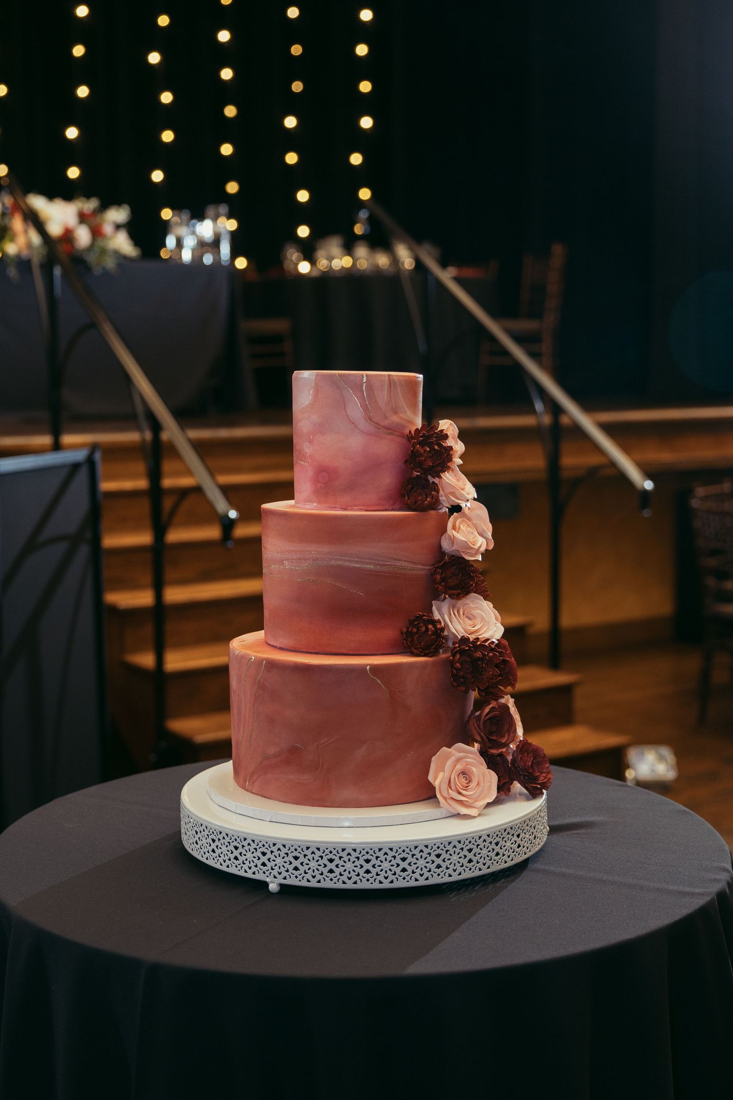 Three-tier pink marble wedding cake with burgundy and blush roses on a white stand in an elegant venue.