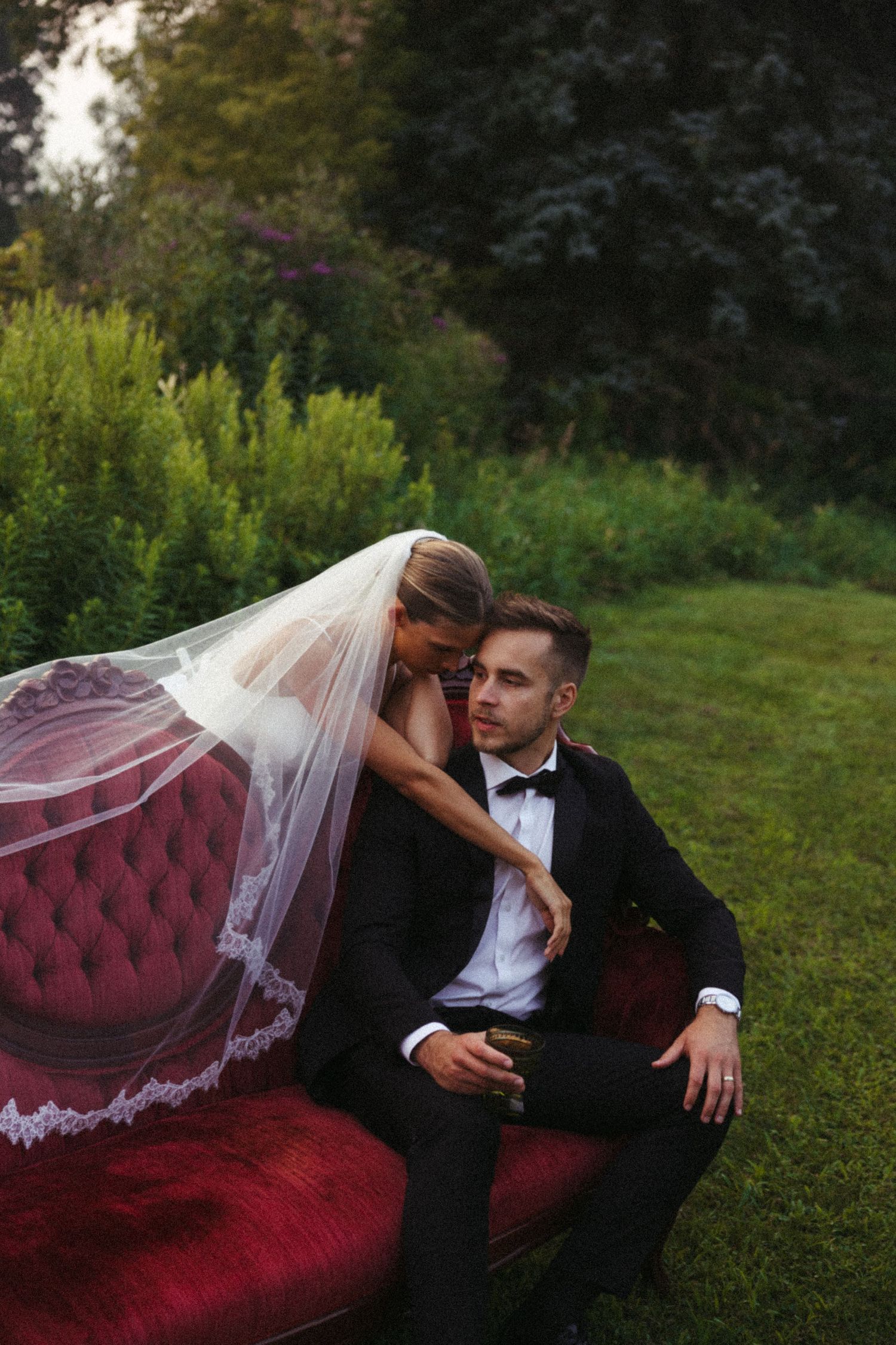 Romantic wedding photo on a red velvet couch outdoors featuring a bride in white veil and groom in black tuxedo.