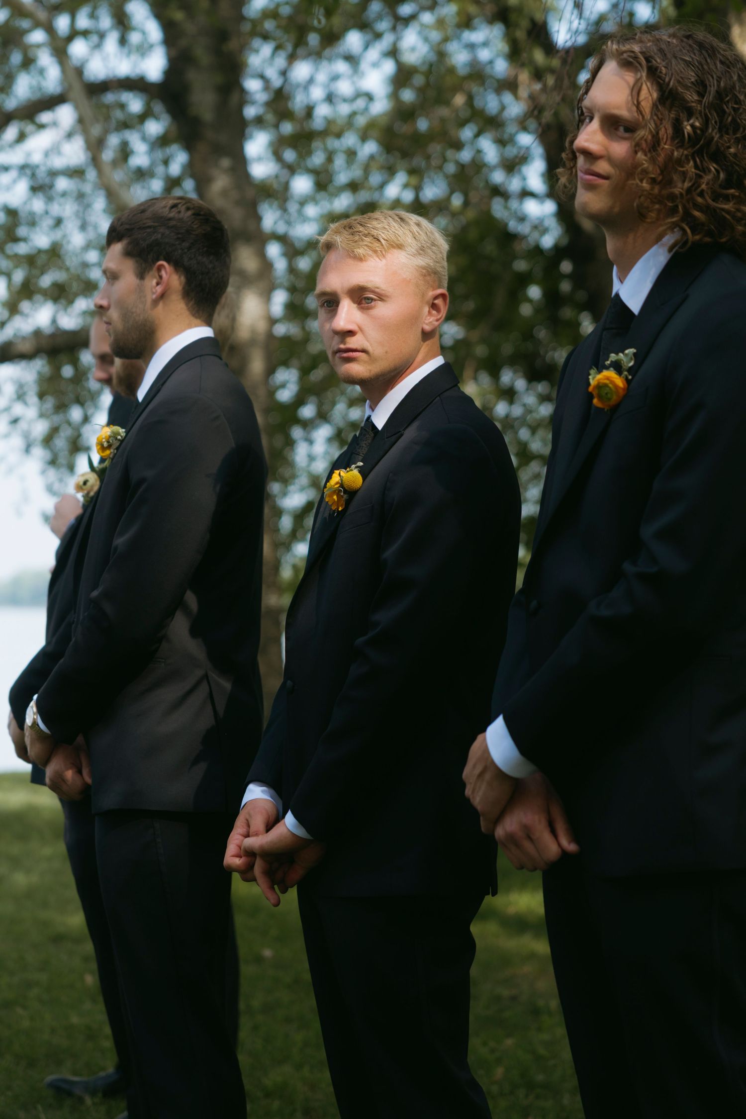 Groomsmen wearing black suits with yellow boutonnieres stand in line at an outdoor wedding ceremony.