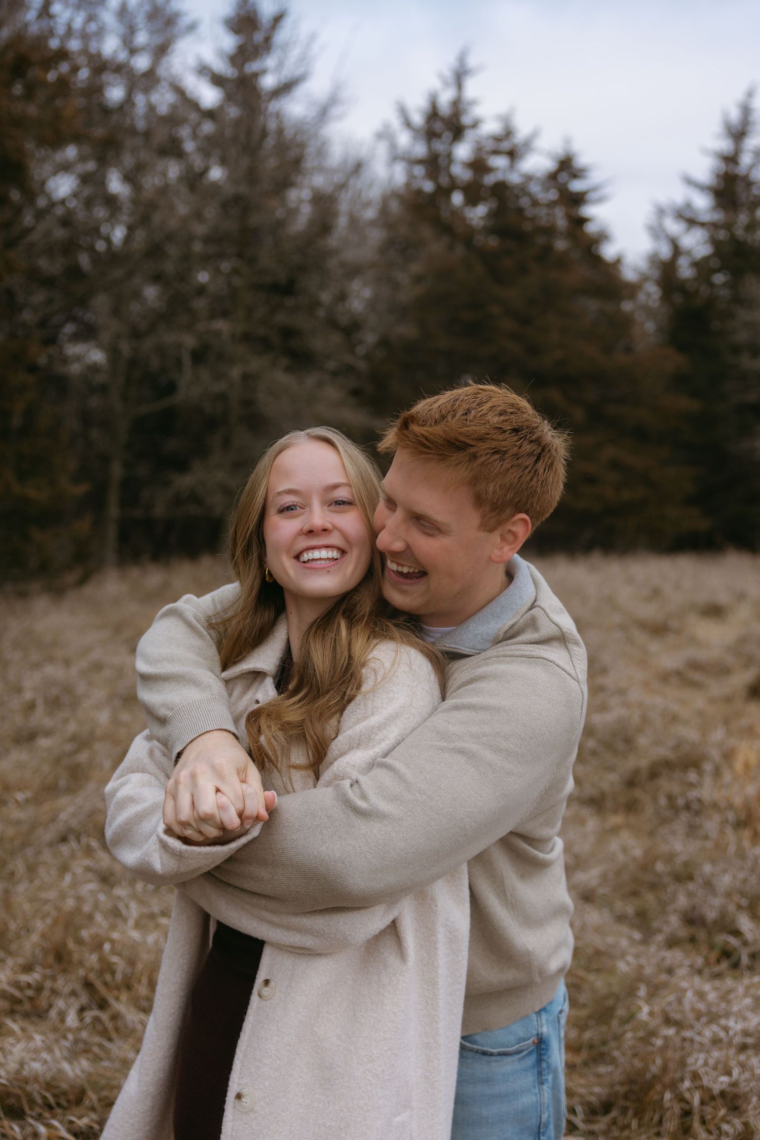 A couple in light neutral sweaters embraces and laughs together in a natural outdoor setting with evergreen trees.